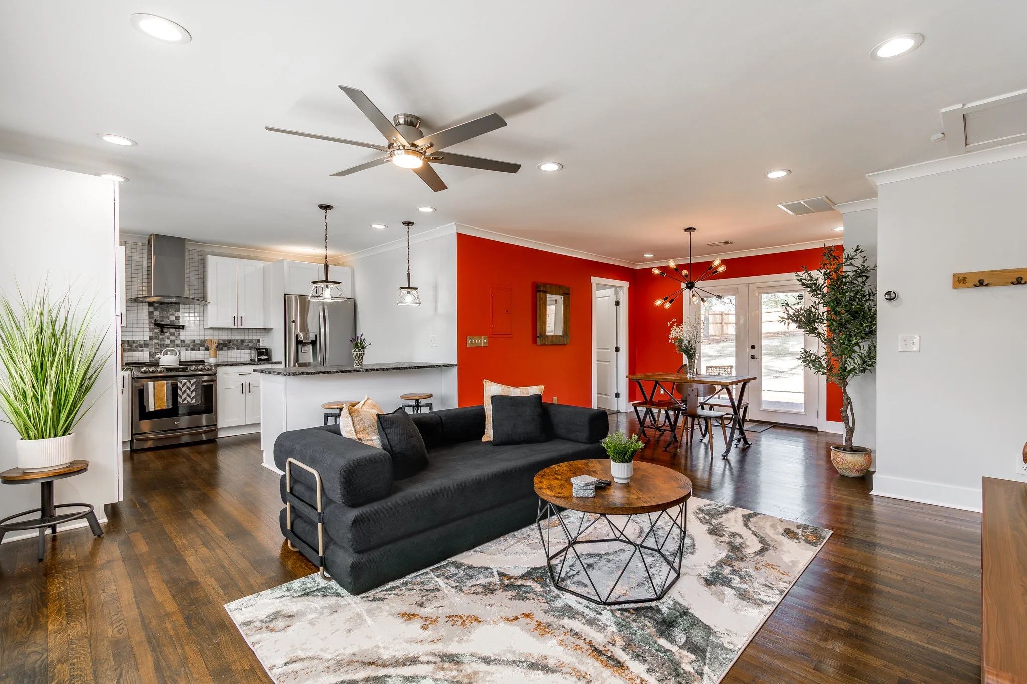 Open-concept living room and kitchen with white cabinets, stainless steel appliances, a black sofa, wooden coffee table, and dining table with chairs, with a vibrant orange accent wall.