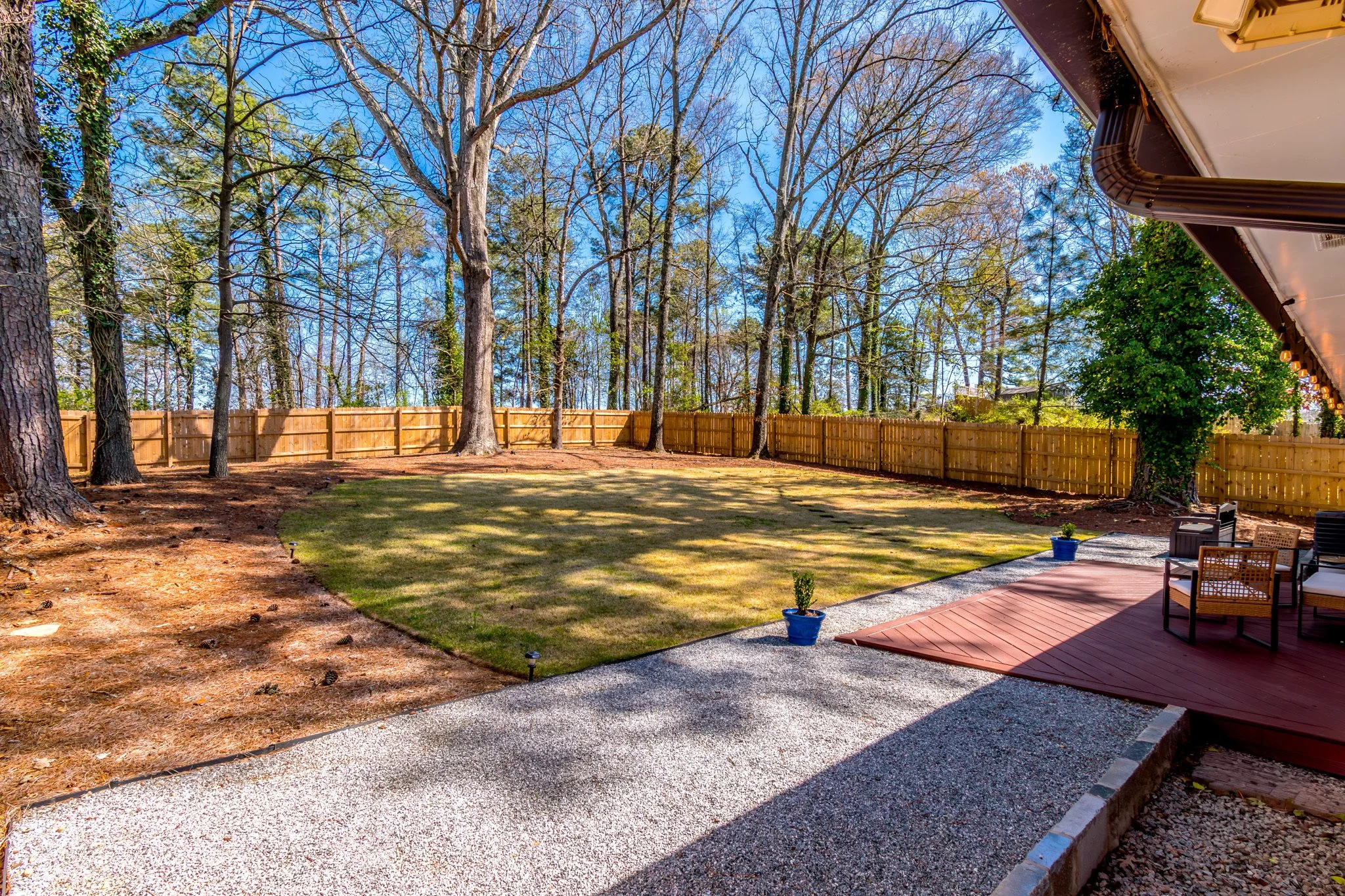 Backyard with a wooden privacy fence, tall trees, a grassy lawn, and a deck with outdoor furniture and potted plants, on a sunny day.