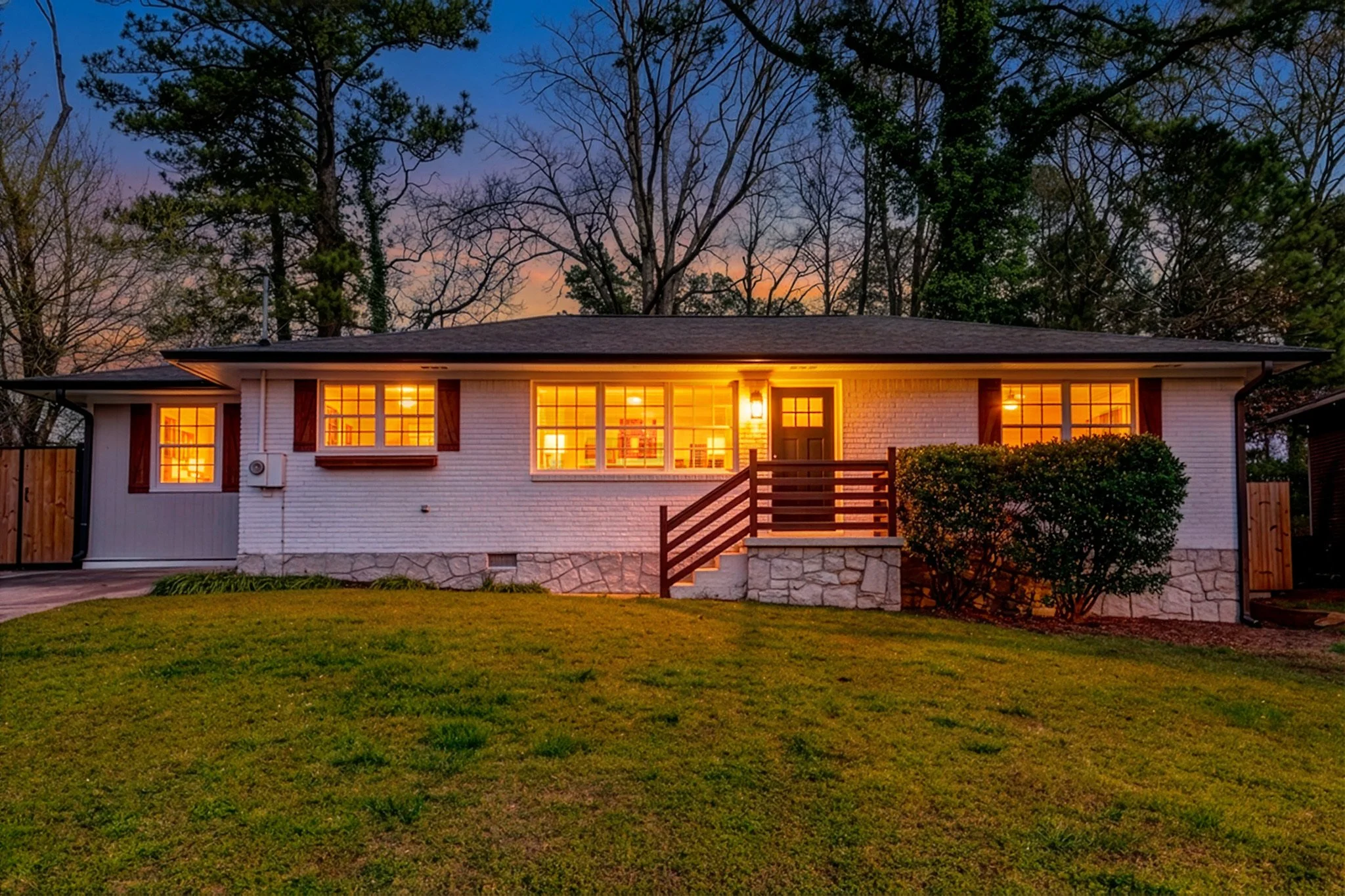 A single-story house with illuminated windows at dusk, surrounded by a green lawn and tall trees.