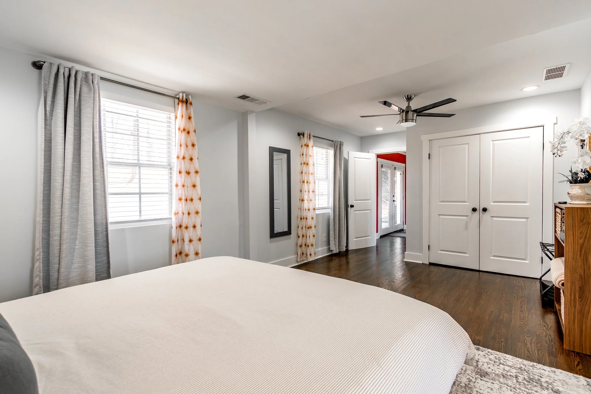 Bright bedroom with white walls, dark wood flooring, large bed with white bedding, two windows with white blinds and patterned curtains, ceiling fan, white closet doors, and a hallway leading to a red accent wall and glass sliding door.