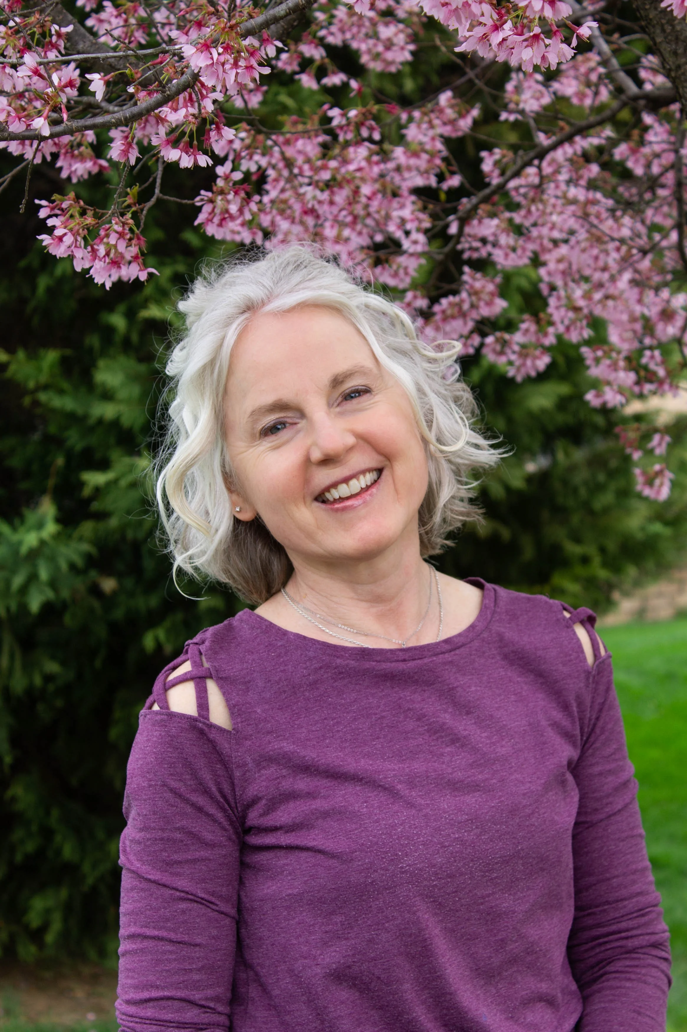 A woman with short, curly gray hair smiling outdoors in front of pink cherry blossom trees, wearing a purple top with shoulder cutouts.