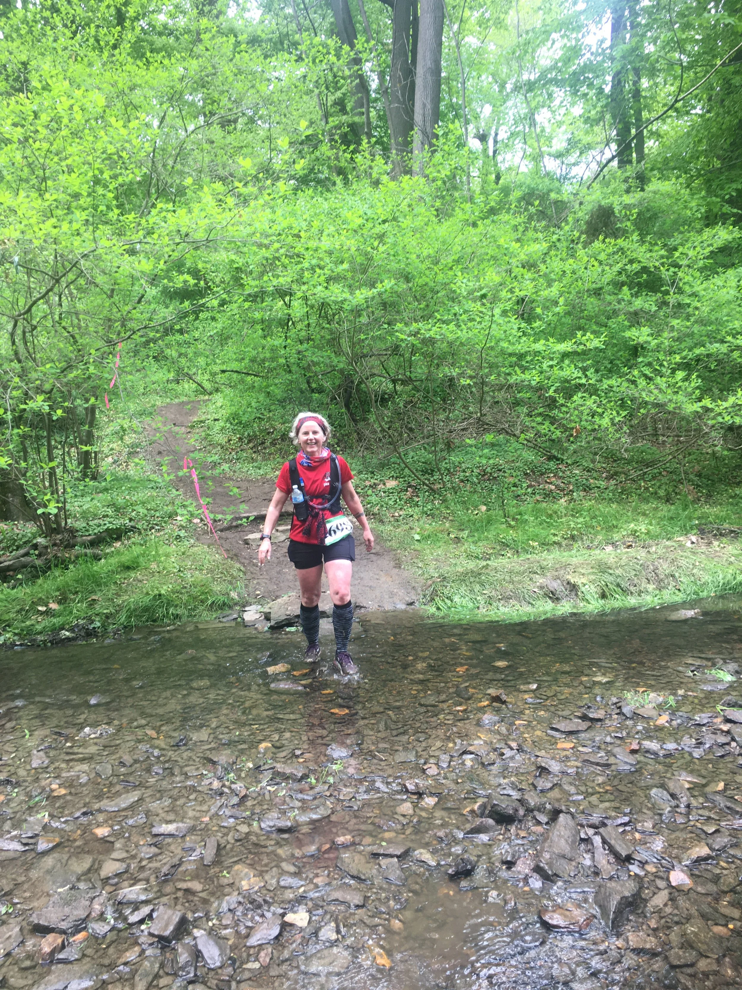 A smiling woman in red shirt and black shorts crossing a shallow creek in the woods during a trail race.