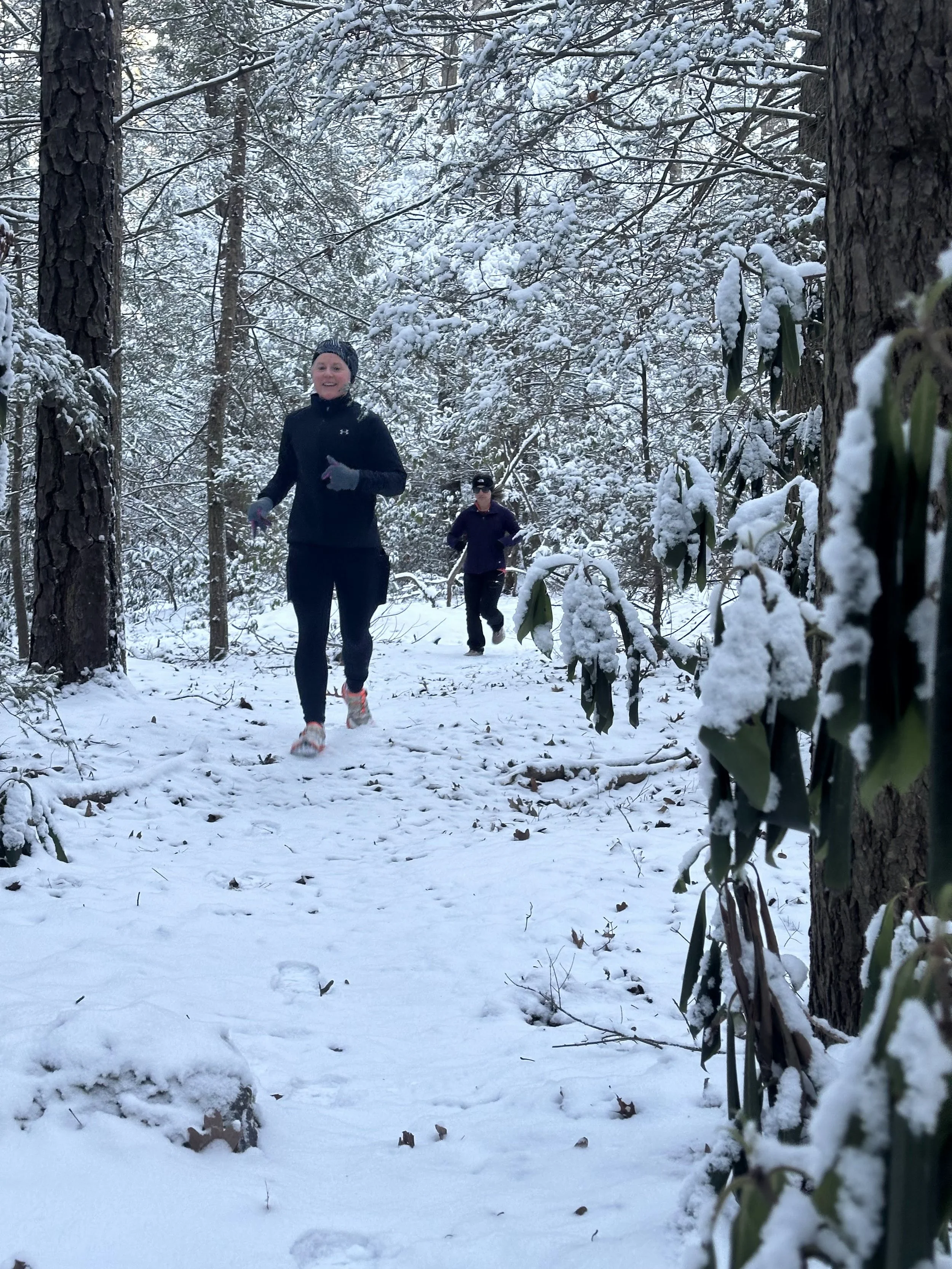 Two women running on a snow-covered trail in a forest with snow-dusted trees and bushes.