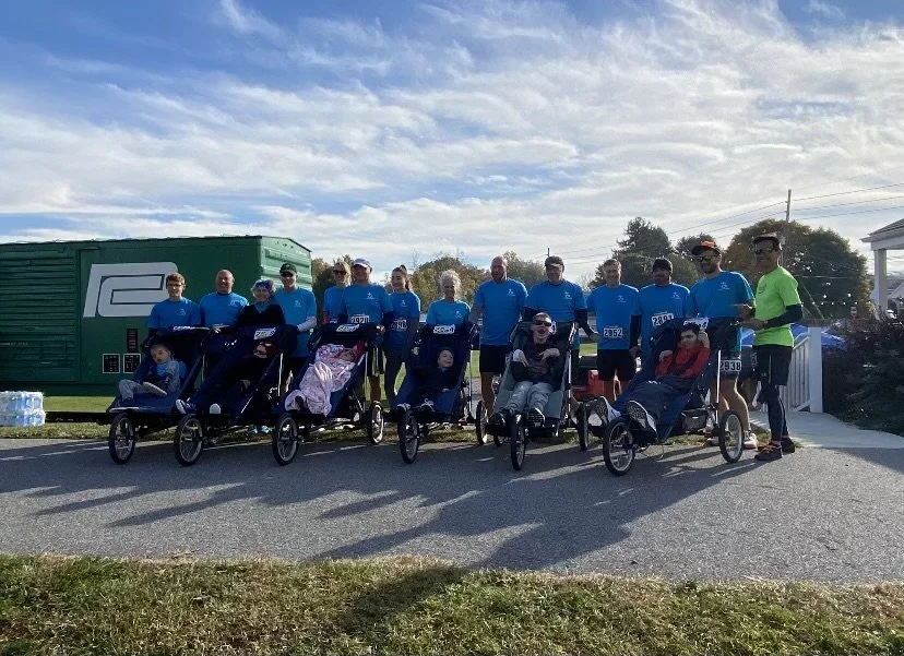 Group of people standing outdoors with children in recumbent tricycles, possibly after a race or event, under a partly cloudy sky.