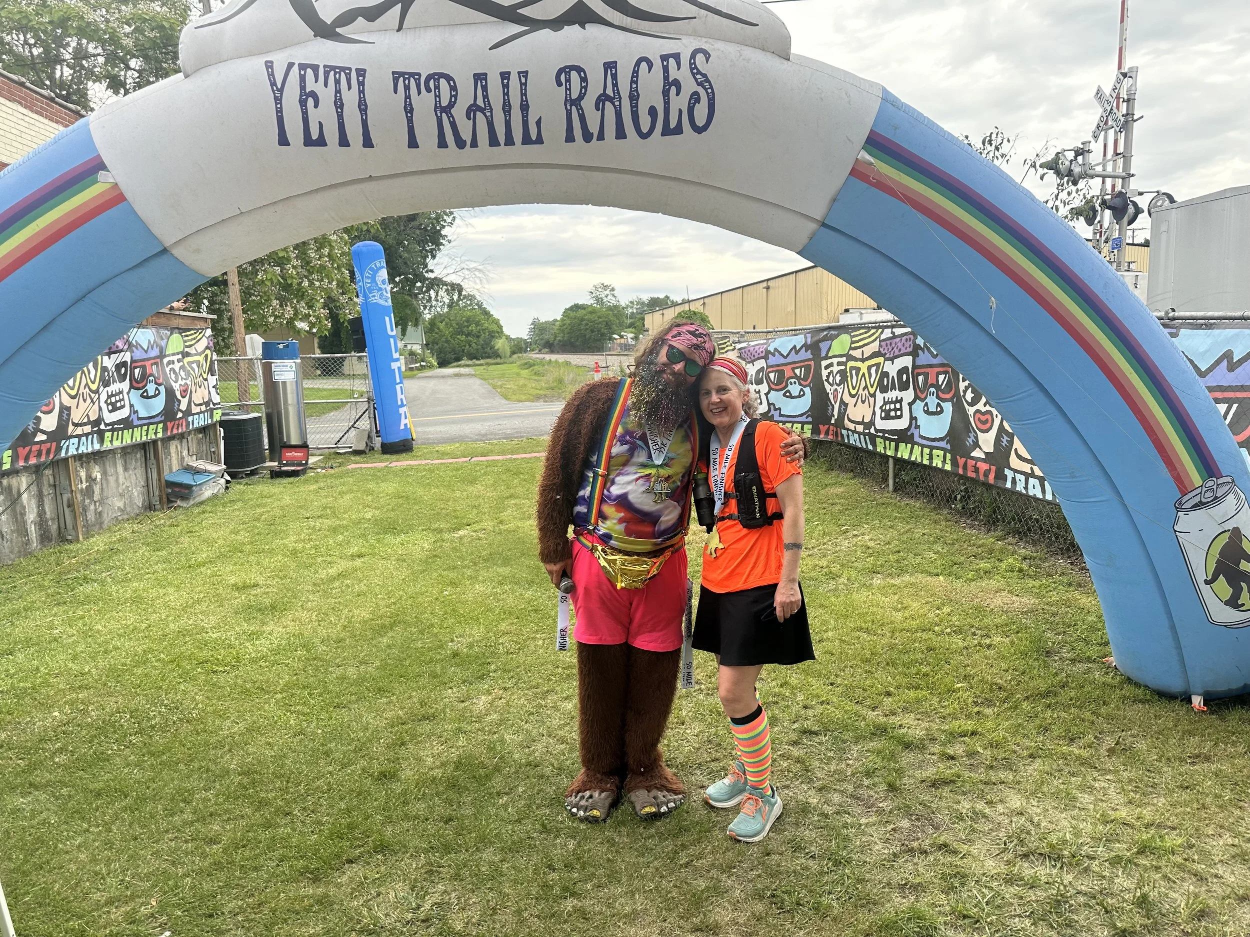 A person with long, curly hair dressed as a yeti and a woman wearing bright orange gaming attire standing together under an inflatable arch that reads 'Yeti Trail Races', with a colorful rainbow design, at an outdoor trail race event.
