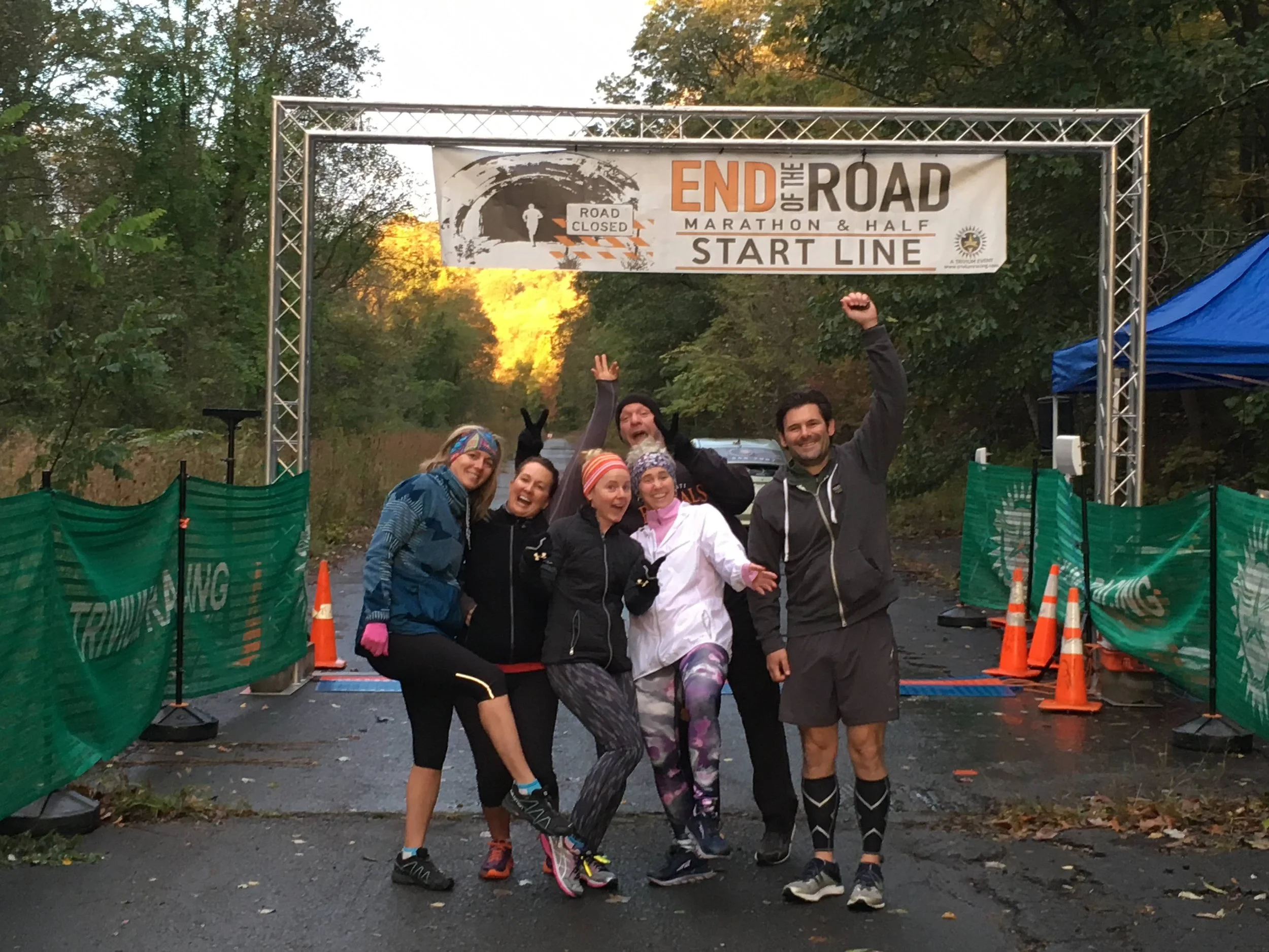 A group of six runners celebrating at the finish line of a marathon or half marathon, standing under a banner that reads 'End of the Road Marathon & Half Start Line', with trees and a road in the background.