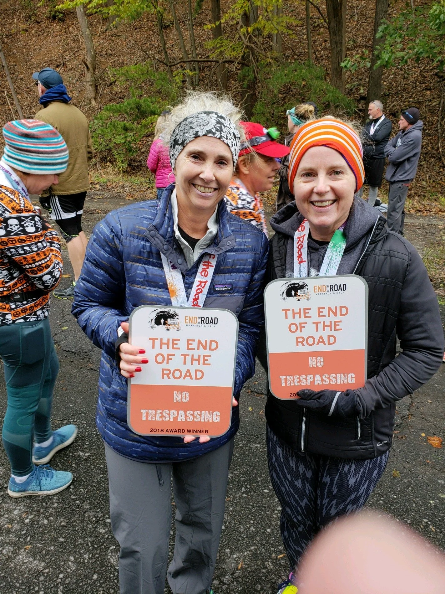 Two women smiling at the camera, holding awards that say 'The End of the Road, No Trespassing,' during a marathon event, with other runners and a wooded area in the background.