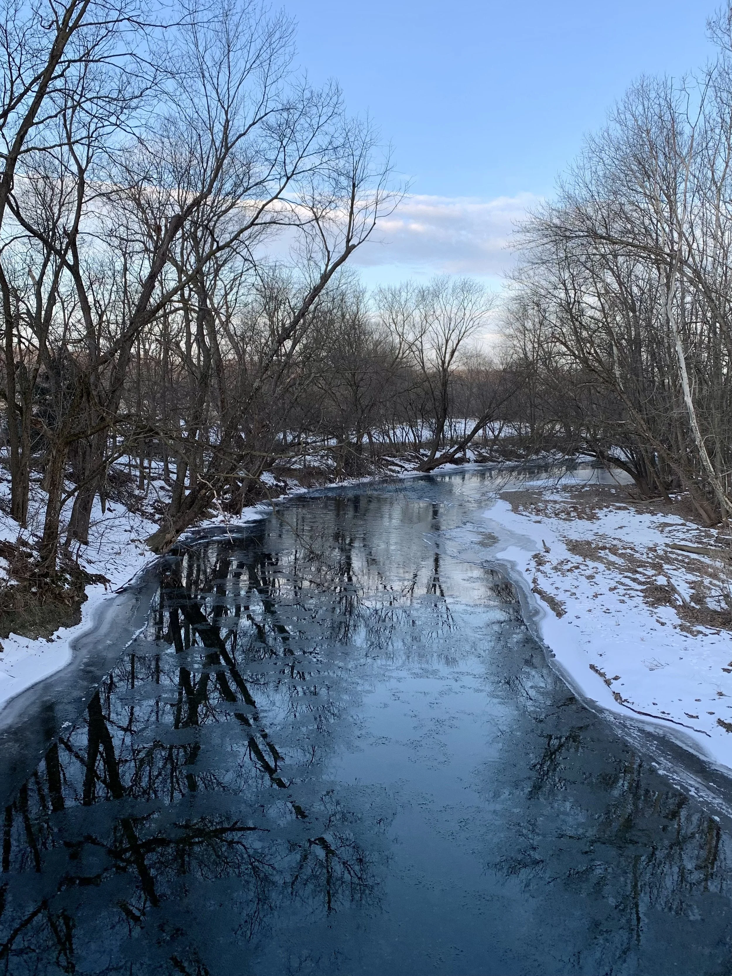 A partially frozen river flowing through a snow-covered landscape with leafless trees on both sides under a blue sky with scattered clouds.