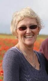 Smiling woman with short blonde hair wearing sunglasses and a purple sweater, outdoors with a field of orange flowers in the background.