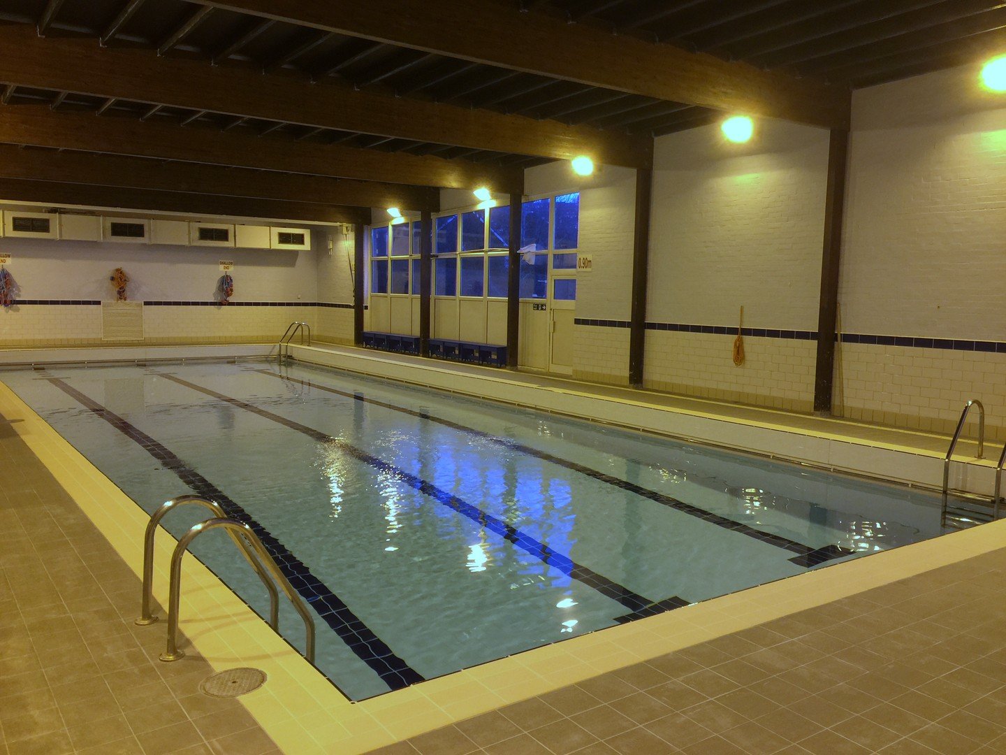Indoor swimming pool with lanes, ladders, and overhead lighting, enclosed by tiled walls and a wooden ceiling.