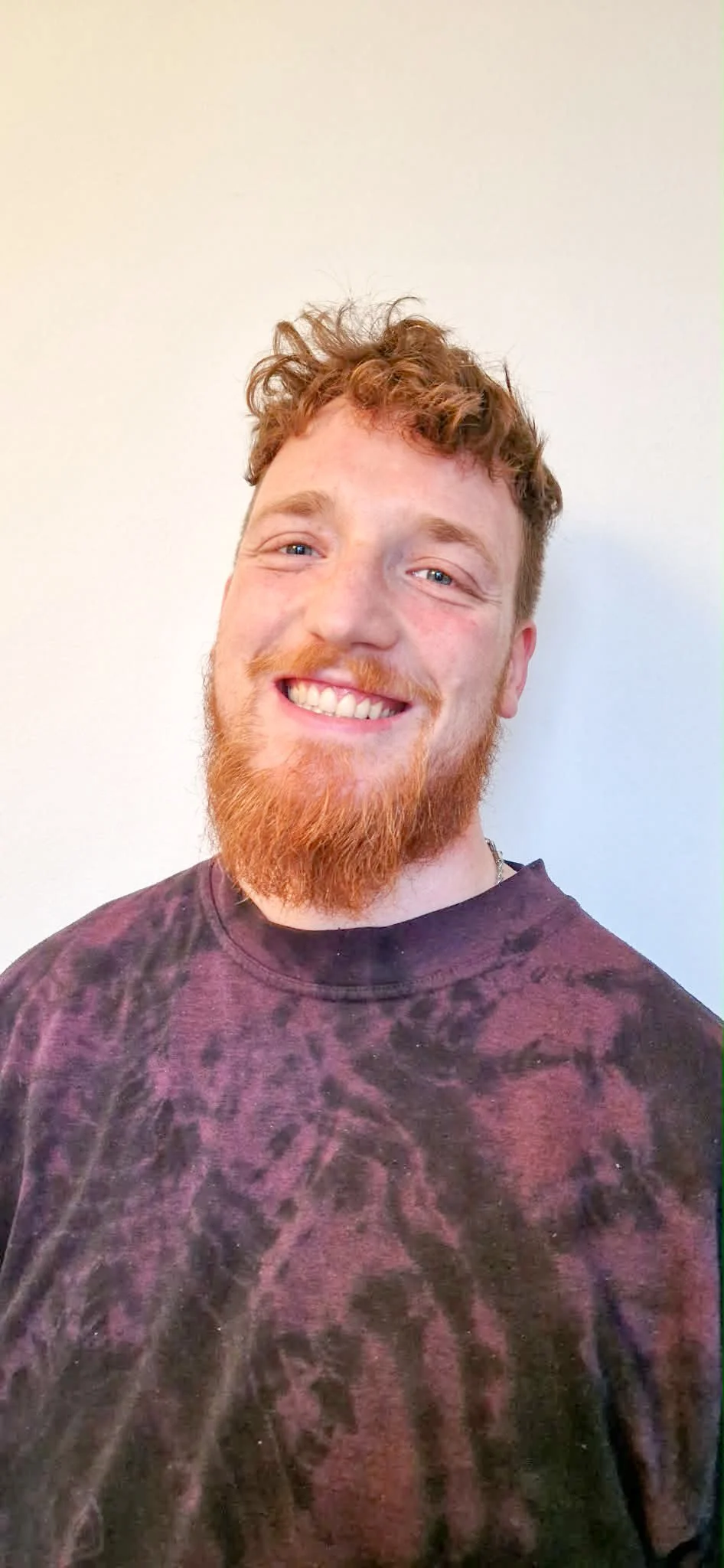 Young man with curly ginger hair and beard smiling, wearing a black and purple tie-dye shirt, standing against a plain white wall.