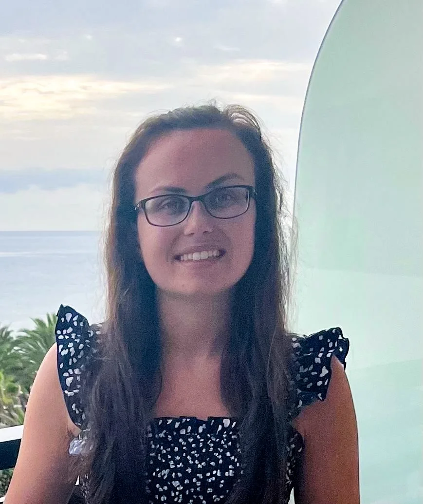 A woman with long dark hair, glasses, and a black dress with white polka dots smiling outdoors near a body of water, with trees and a cloudy sky in the background.