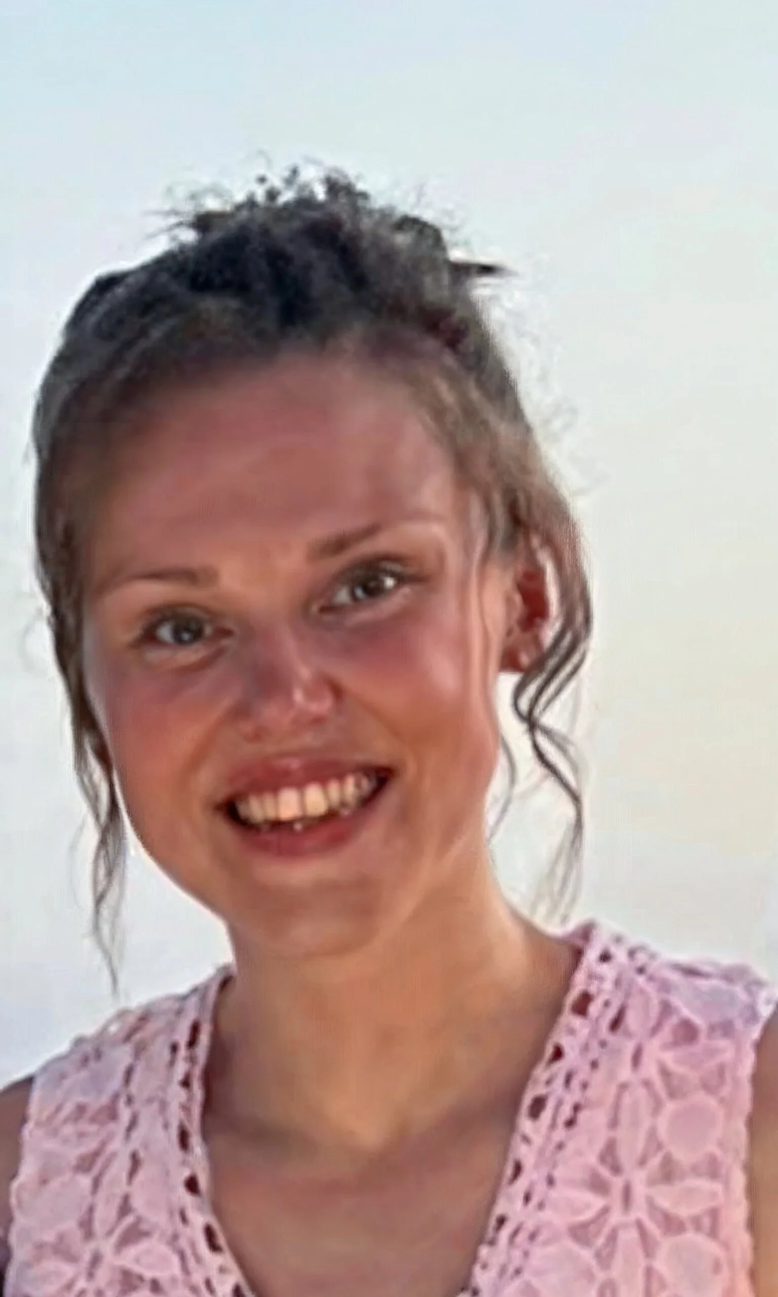 A smiling woman with curly hair wearing a pink lace dress against a plain light background.