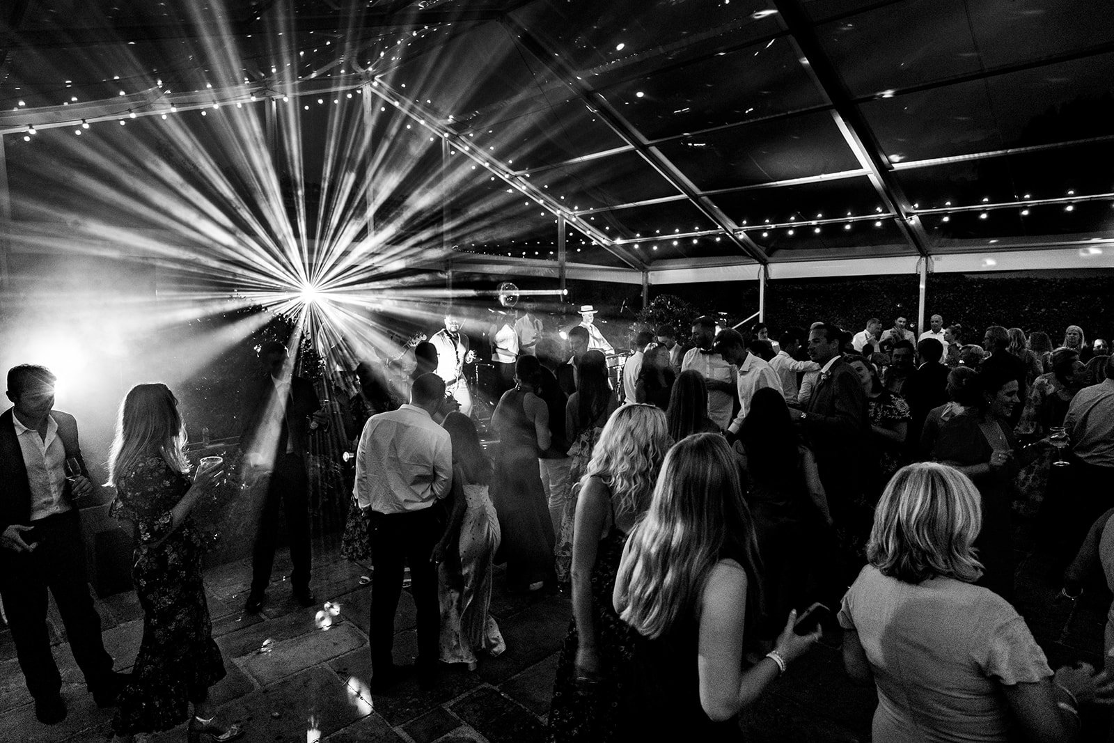 Black and white photo of a lively indoor party with many people dancing and socialising under string lights with a band playing live on stage and a large, bright light in the background. The party is in a large glass marquee and everyone's having fun