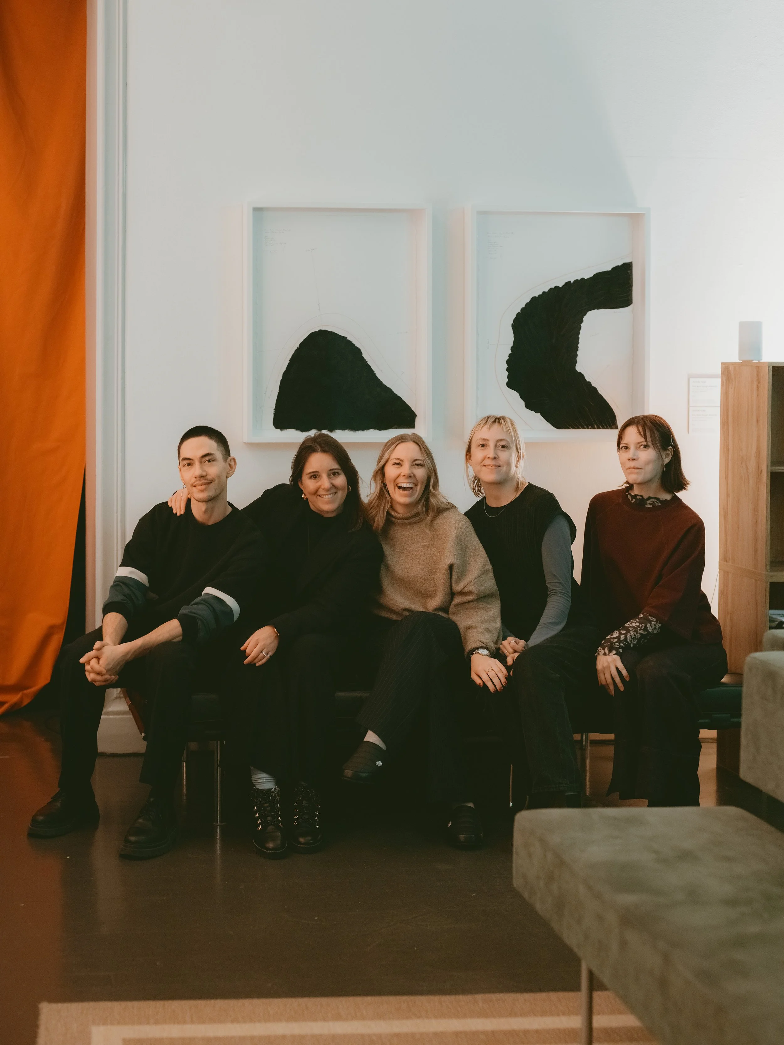 Group of five colleagues sitting on a black bench in a venue, smiling and posing for the photo, with two abstract artworks hanging on the white wall behind them.
