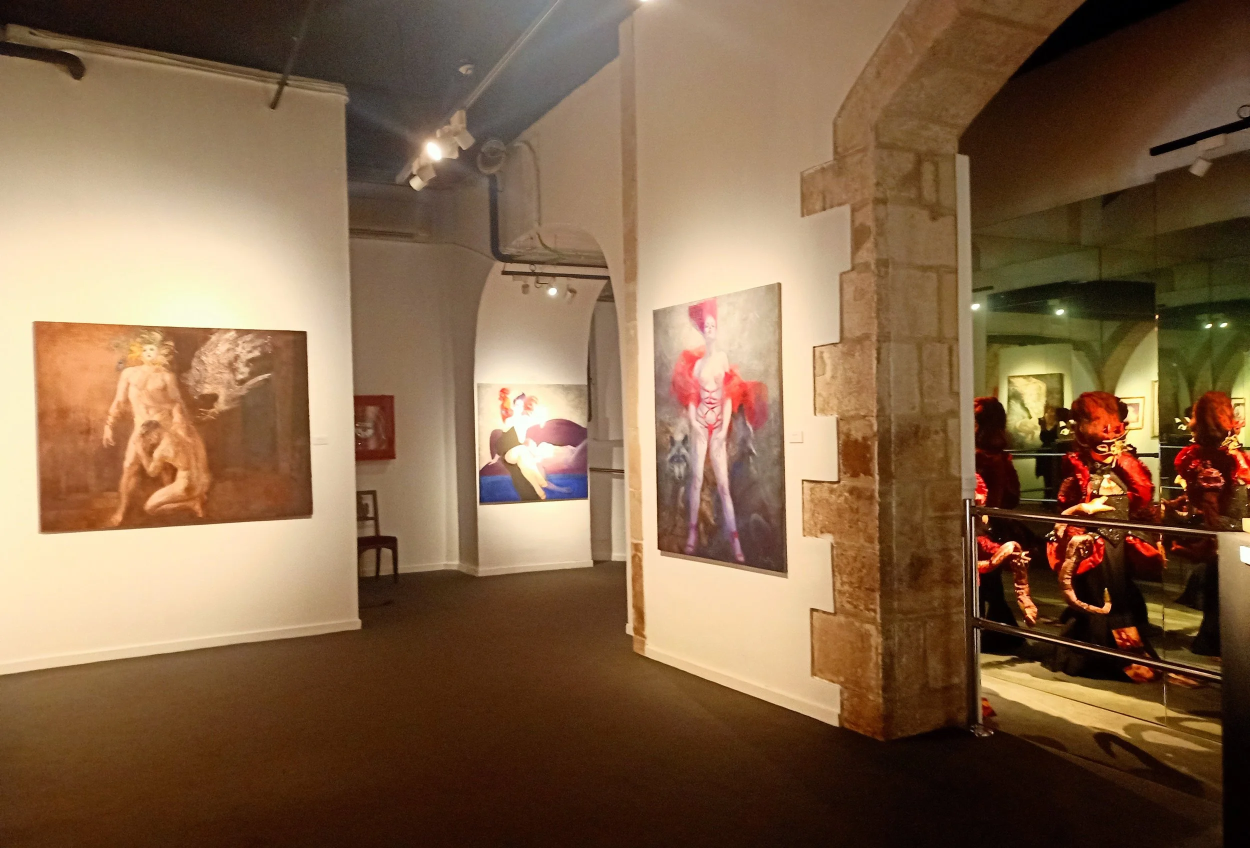 Art gallery with paintings on white walls, showing abstract and fantasy figures; some visitors in the background observe the artwork.