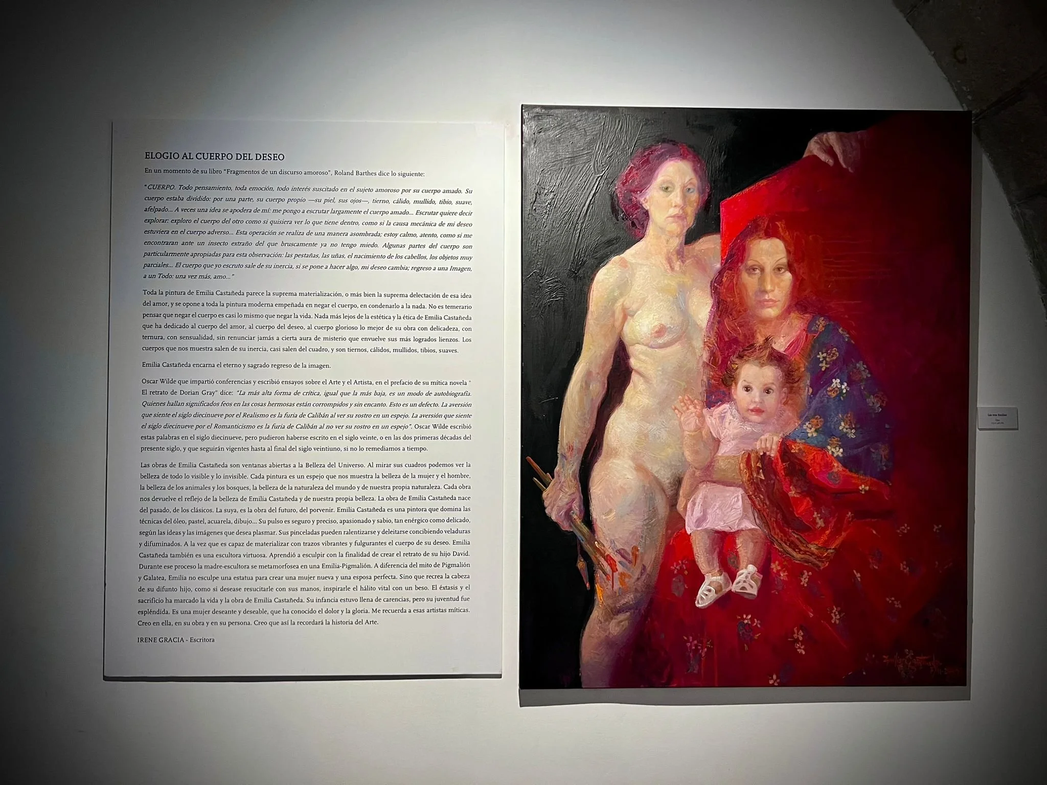 Painting of a woman with red hair holding a toddler, another woman with pink hair, and a young girl, displayed at an art gallery alongside a descriptive text panel.