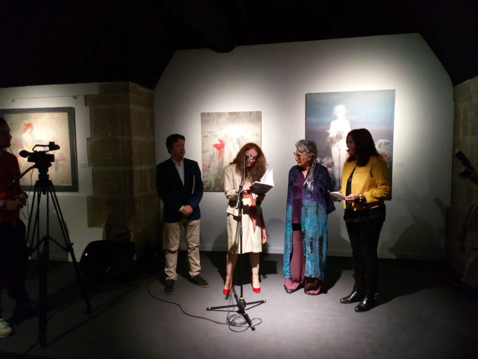 People gathered in an art gallery for an event or presentation, with paintings displayed on the wall behind them. A woman in beige is speaking into a microphone while reading from a book, surrounded by others.