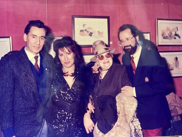 Group of five people posing together at an art gallery, with framed paintings on a red wall behind them.