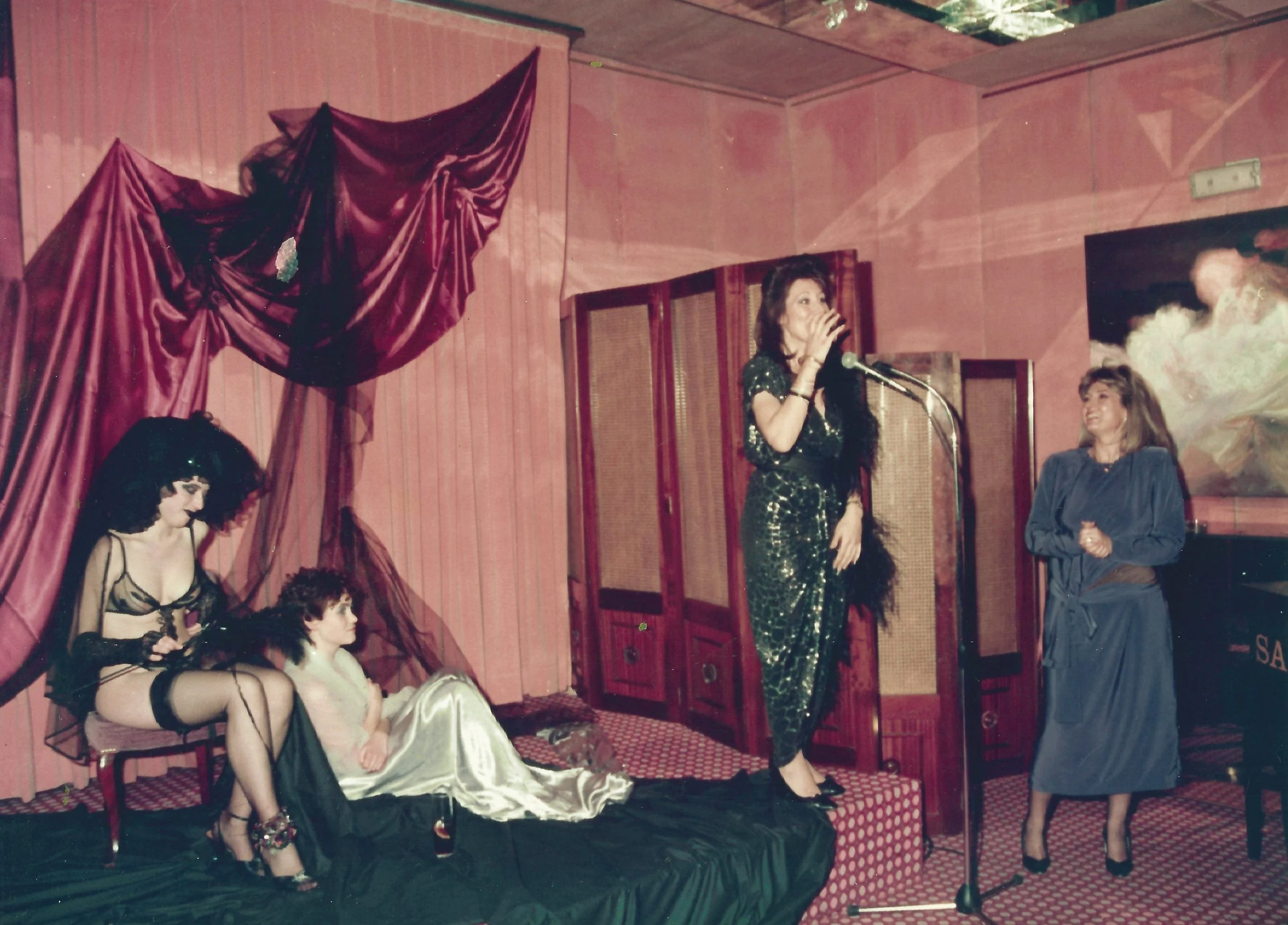 Women dressed in vintage lingerie and costumes sitting on a couch, with a woman in a sequined dress speaking into a microphone on a stage in front of them. The room has pinkish walls and ornate curtains.