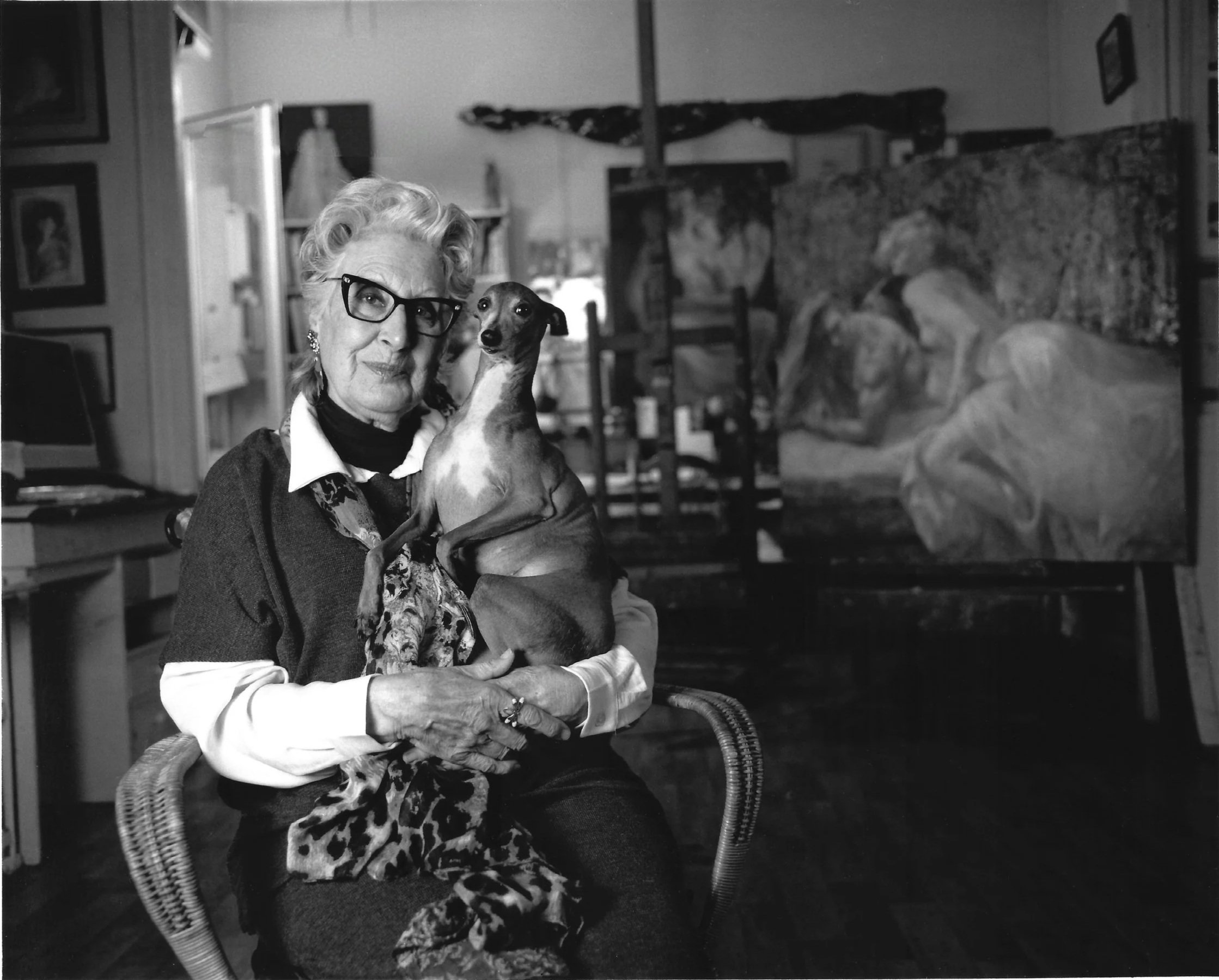 Emilia Castañeda sitting on a wicker chair, holding a Perla her dog with a short coat, in her studio with bookshelves and paintings on the wall.