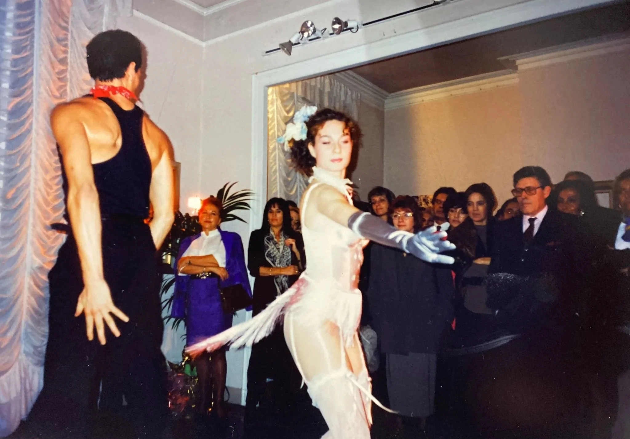 A woman in a fancy white gown with gloves and floral headpiece performing a dance on stage, with a large audience watching.