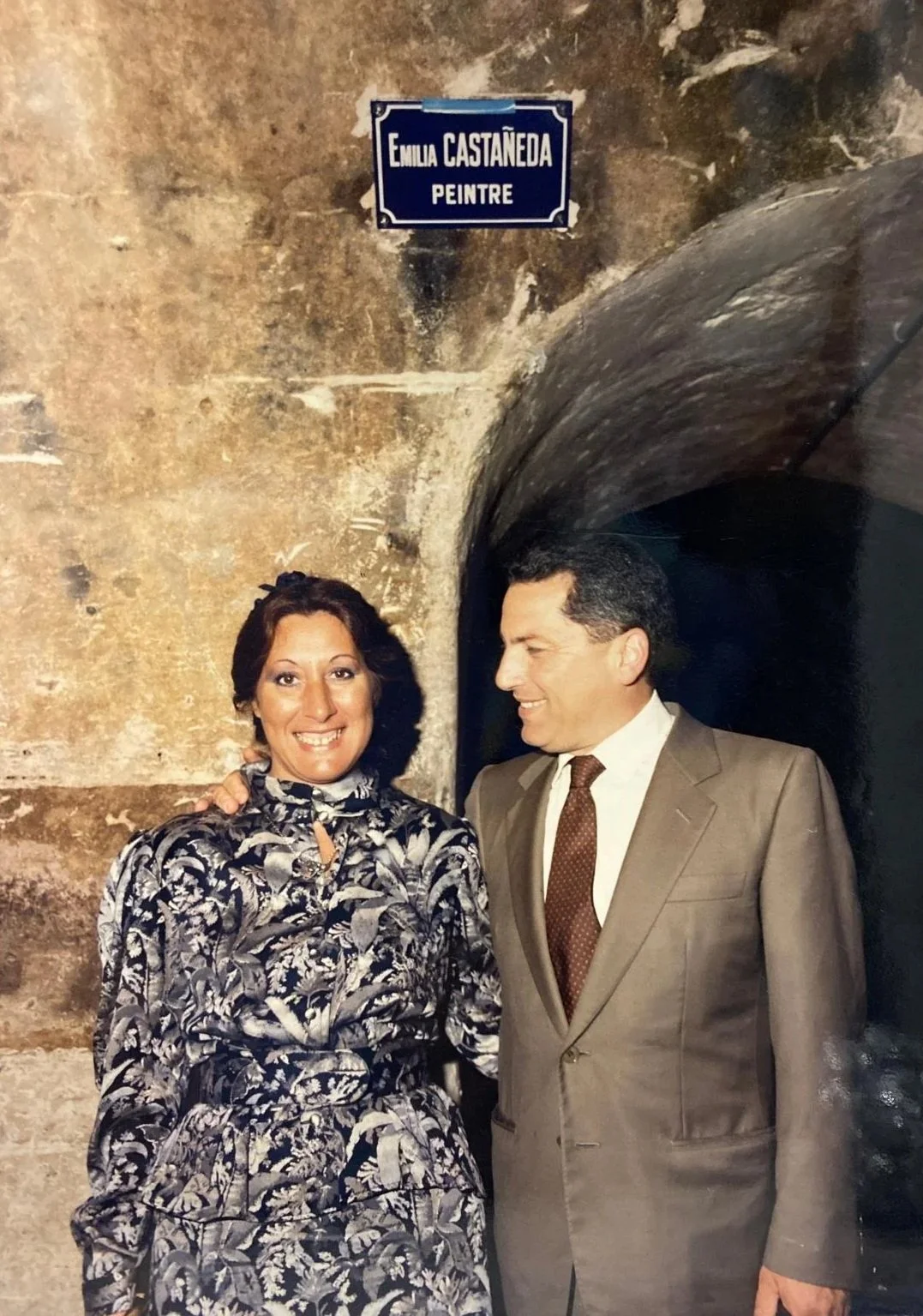 A woman and a man dressed in formal attire standing next to each other, smiling, in front of a stone wall with an archway. There is a blue street sign above them that reads 'Emilia Castañeda Peintre.'