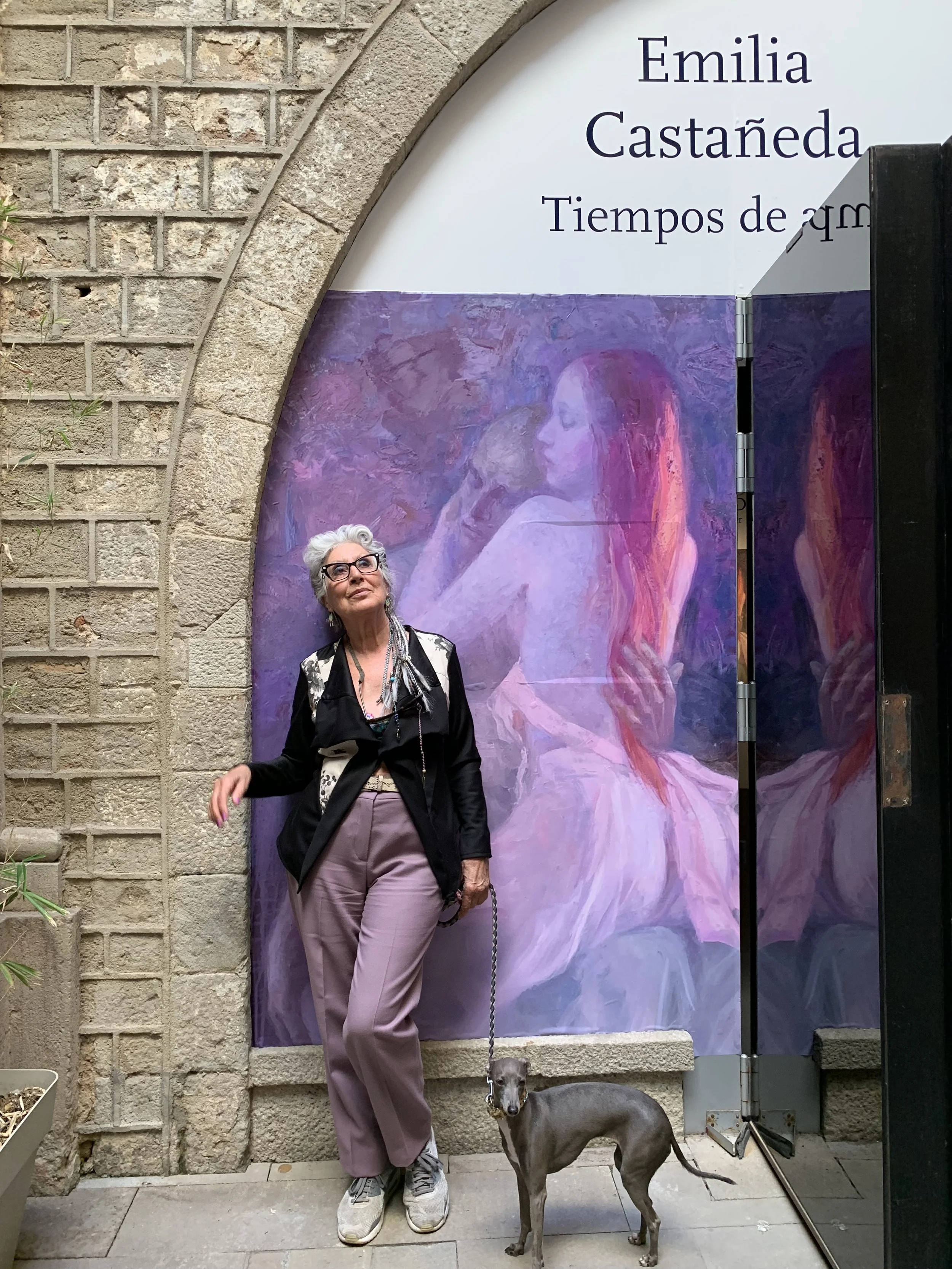 An older woman with gray hair, glasses, and colorful clothing standing next to her gray Italian Greyhound dog in front of an art display at Emilia Castañeda's exhibit. The display features a purple and pink painting of a woman with long hair.