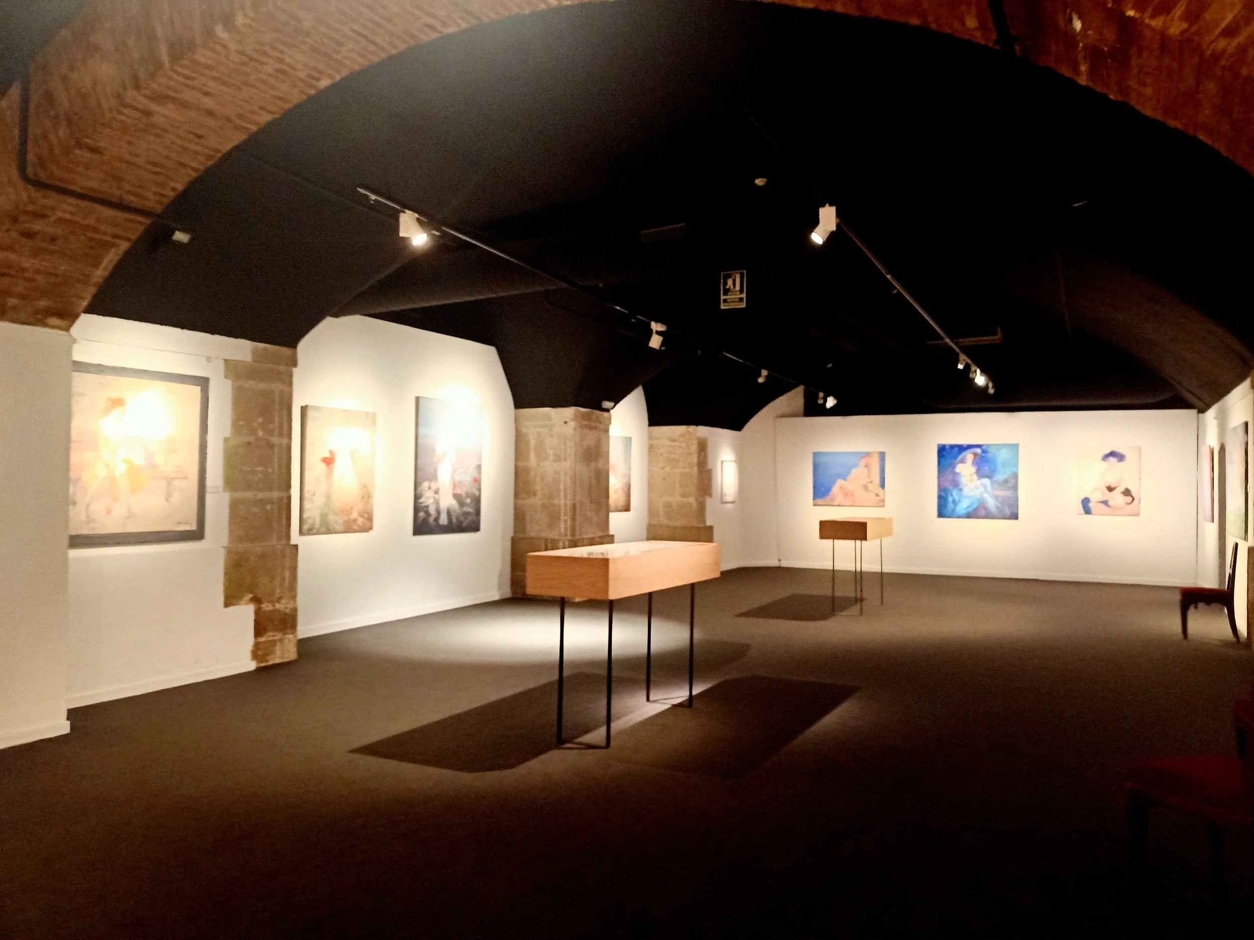 An art gallery with paintings on white walls, a black ceiling with track lighting, and wooden display tables. There are chairs along the walls.