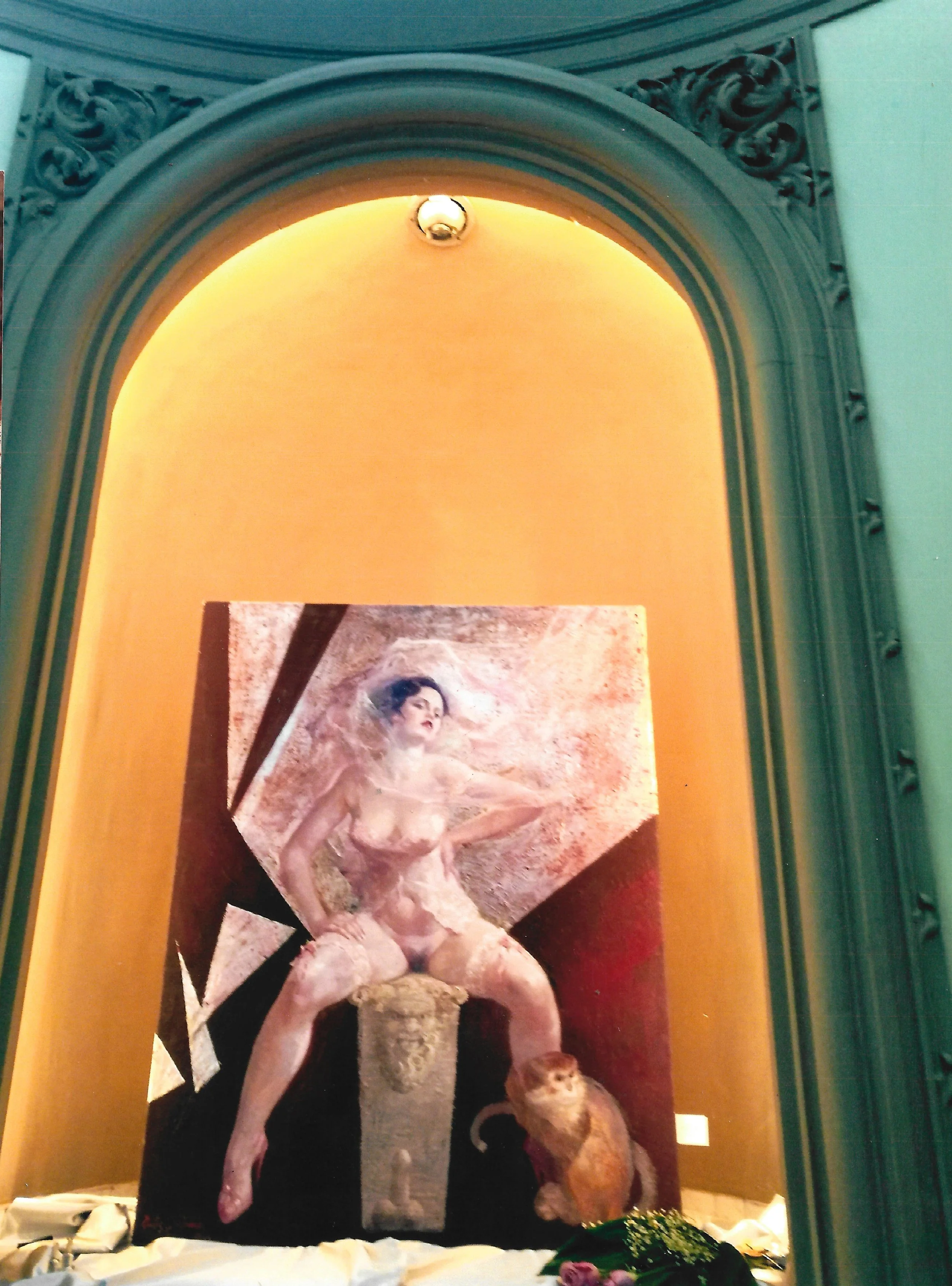 An art piece featuring a nude woman with curly hair, wearing lace stockings, seated on a stage or platform with artwork in the background. The image is displayed on a screen or in a frame with ornate green trim.