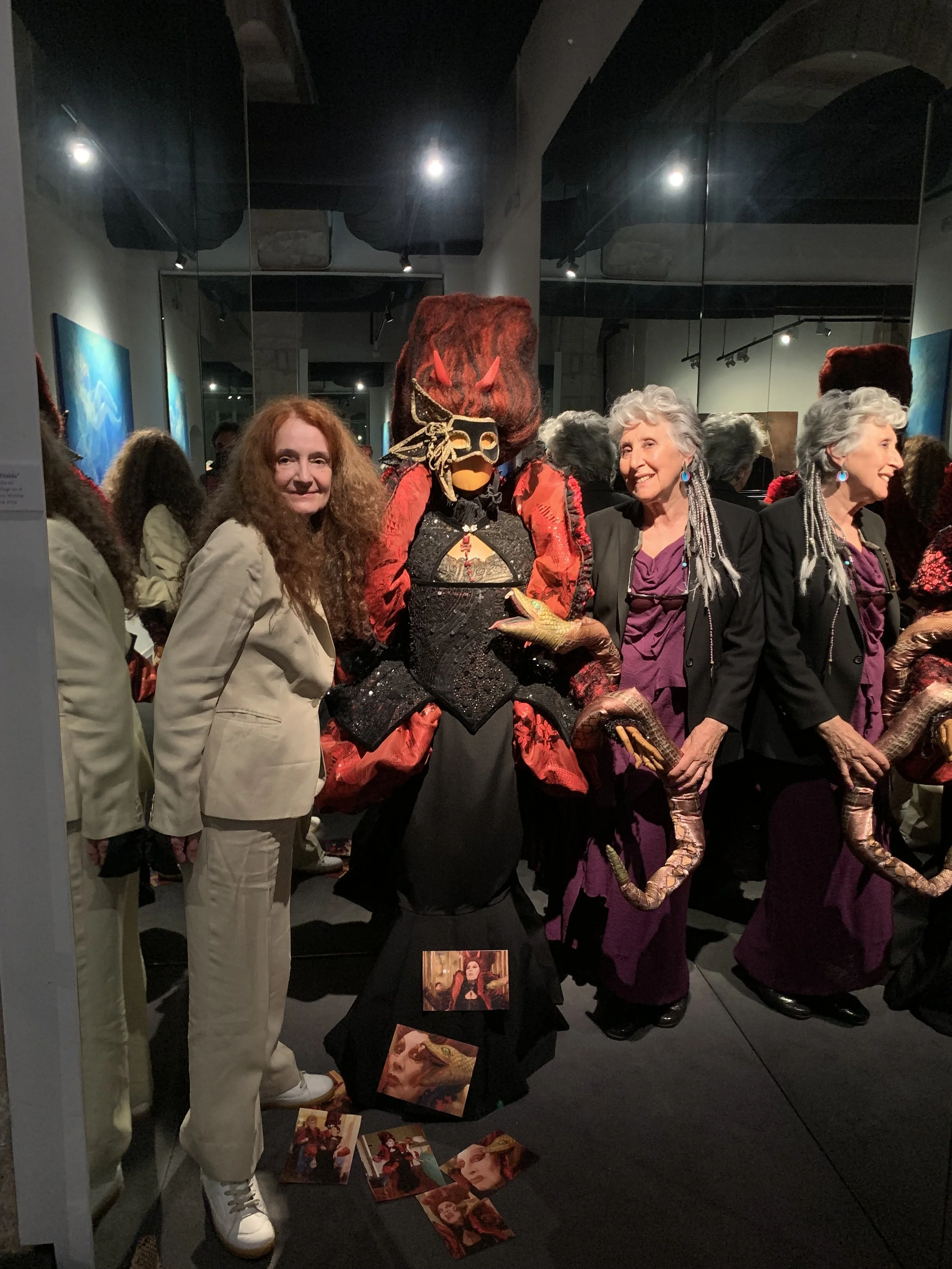 Two women and a large puppet or sculpture resembling a mythical creature with multiple limbs, vibrant red and black clothing, and a fierce face, displayed in an art gallery with reflections in mirrors and artworks on the walls.
