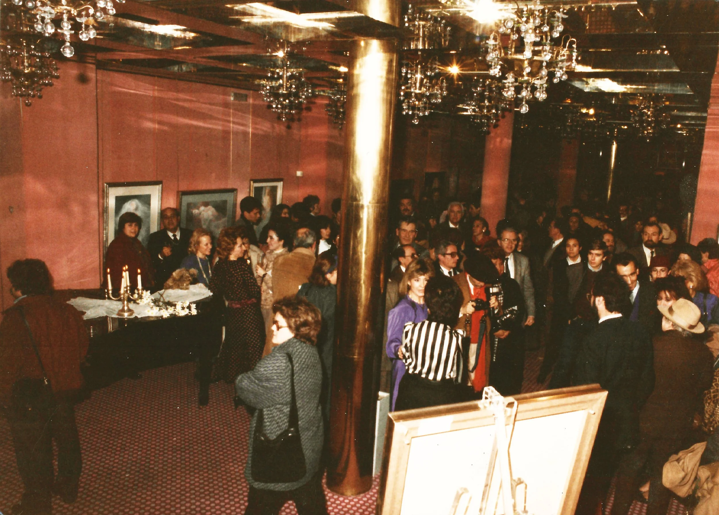 A crowded indoor gathering with people dressed in formal attire, socializing under chandeliers with glass ornaments, with a table decorated for a celebration in the foreground.