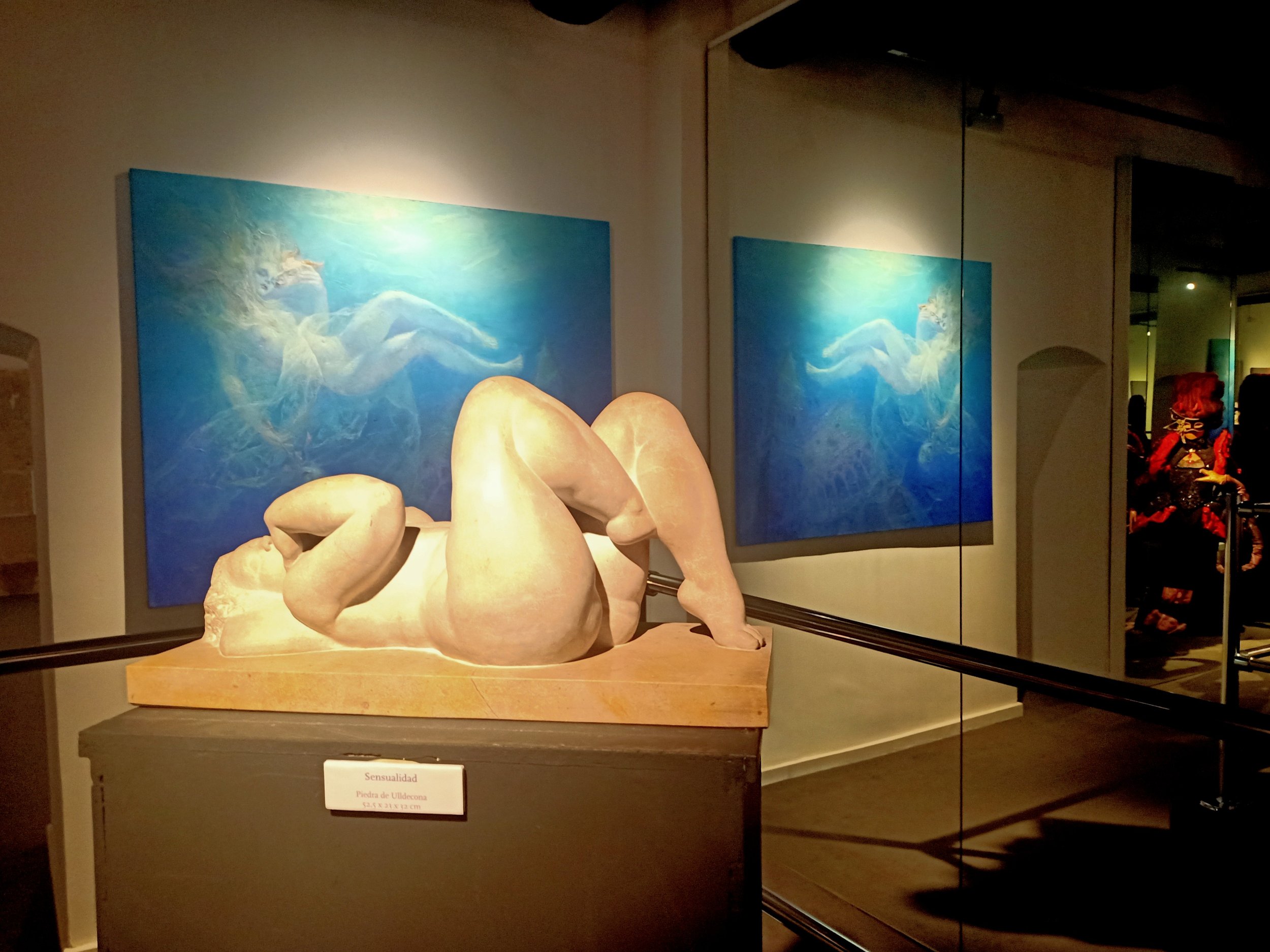 A marble sculpture of a reclining nude woman with legs bent, on display in front of blue-toned paintings of a mythological figure with a horse and wings in an art gallery.