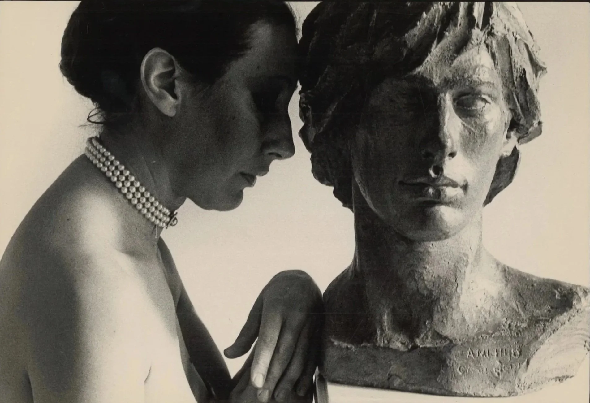 Black and white photo of a woman wearing pearls, with her head close to a bust sculpture of a woman with textured hair.