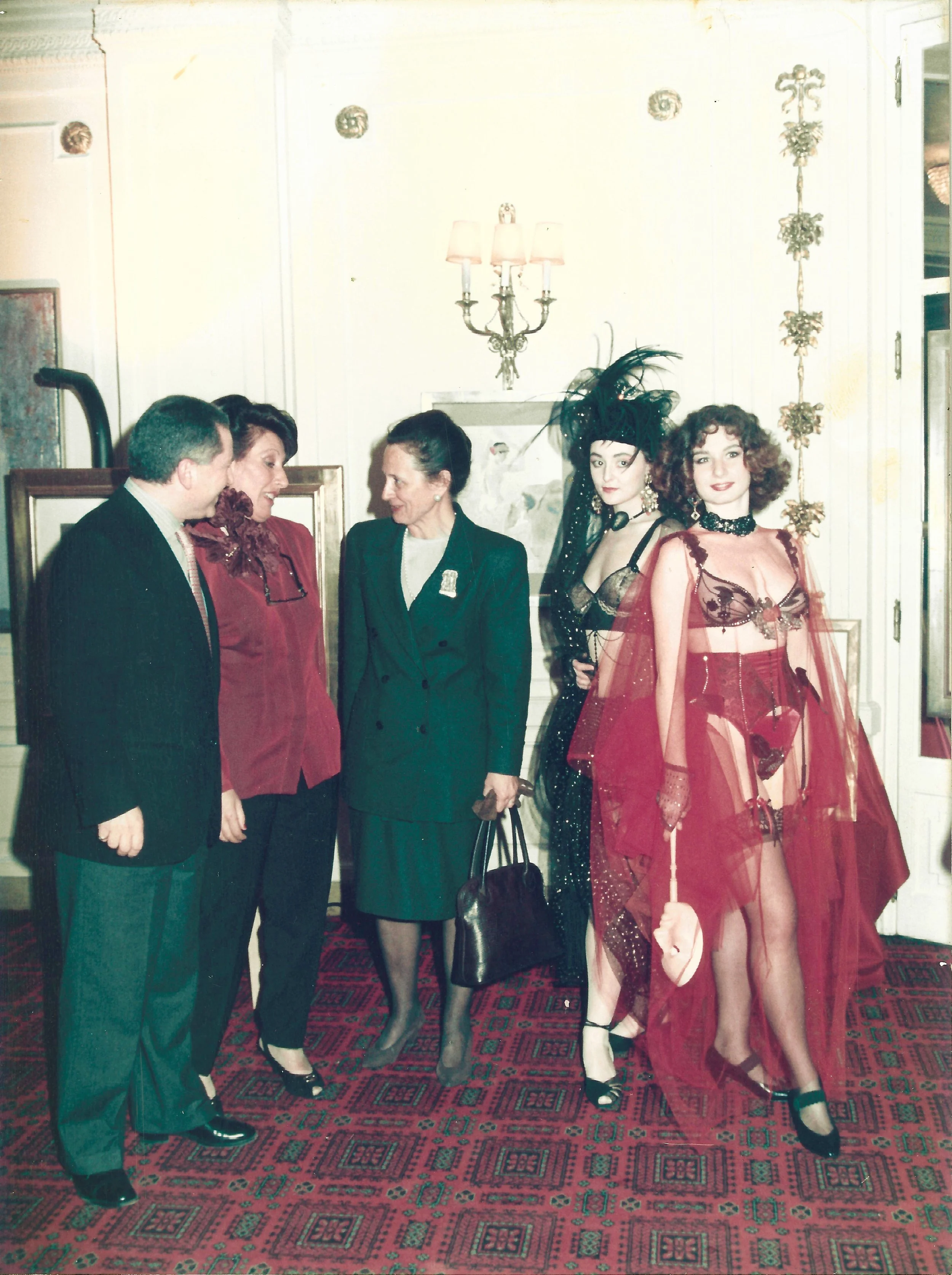 A group of five people standing indoors in a room with patterned carpet and ornate wall decorations. Two women are dressed in elaborate, revealing red outfits with black accents, and three other individuals are dressed in more formal, conservative at