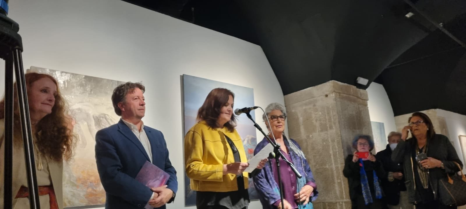 Group of people at an art gallery, some speaking at a microphone, artwork on walls behind them.