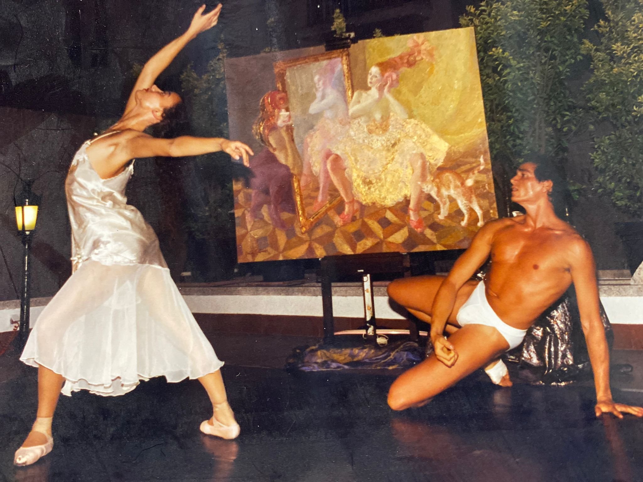 Two dancers performing on stage with a large painting as a backdrop. The dancer on the left is wearing a light-colored dress and ballet shoes, with arms raised and body arched. The dancer on the right is shirtless, wearing white shorts, with one knee