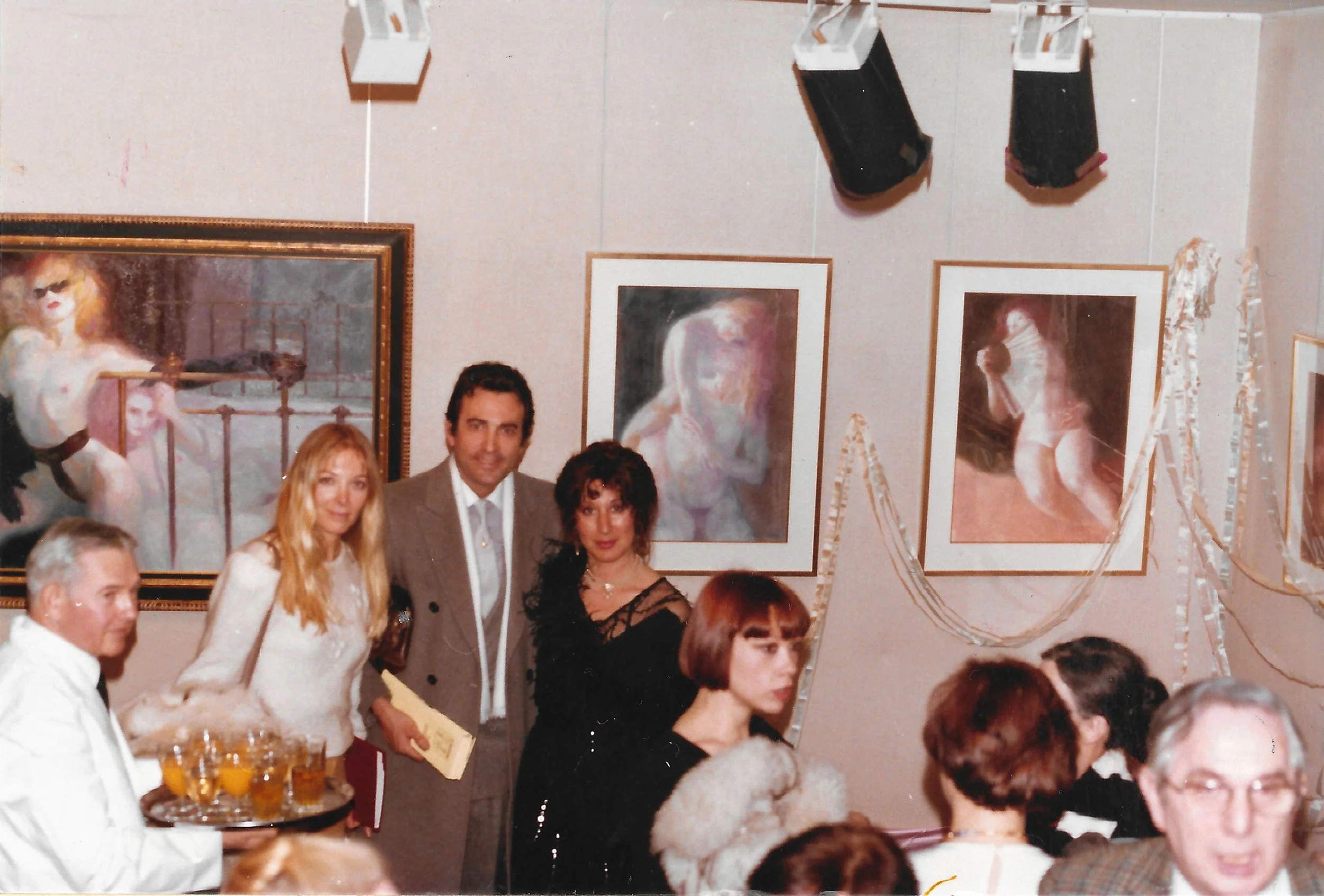 Group of people at a social gathering in an art gallery or museum with paintings on the wall and black speakers hanging from the ceiling.
