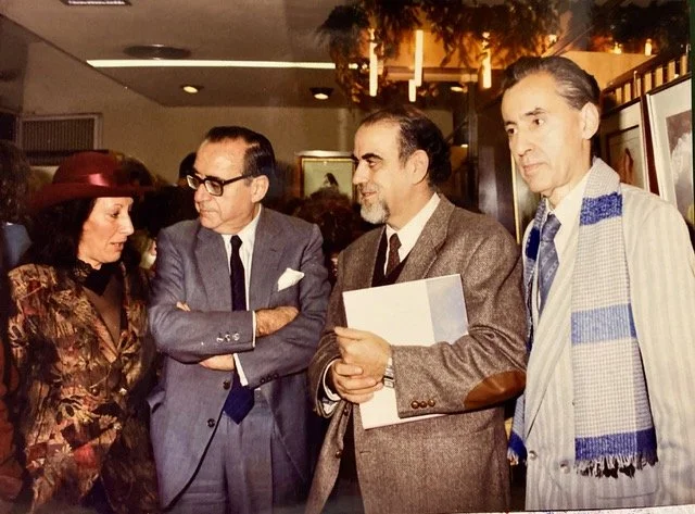 Four people dressed in formal attire, engaged in conversation at an indoor event or gallery.