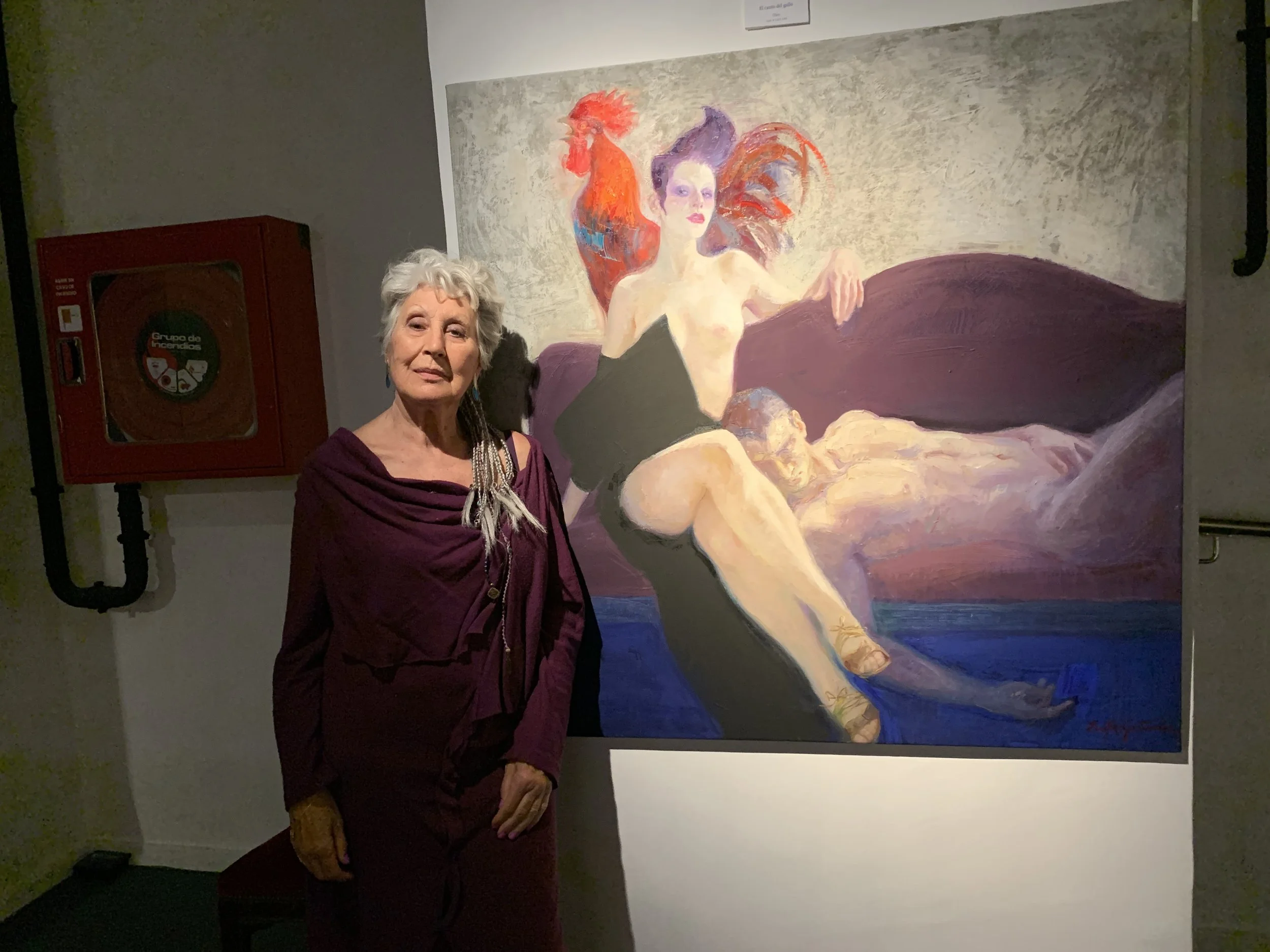 Older woman in a purple dress with a shawl stands in front of a large abstract painting of three women in a reclining scene.