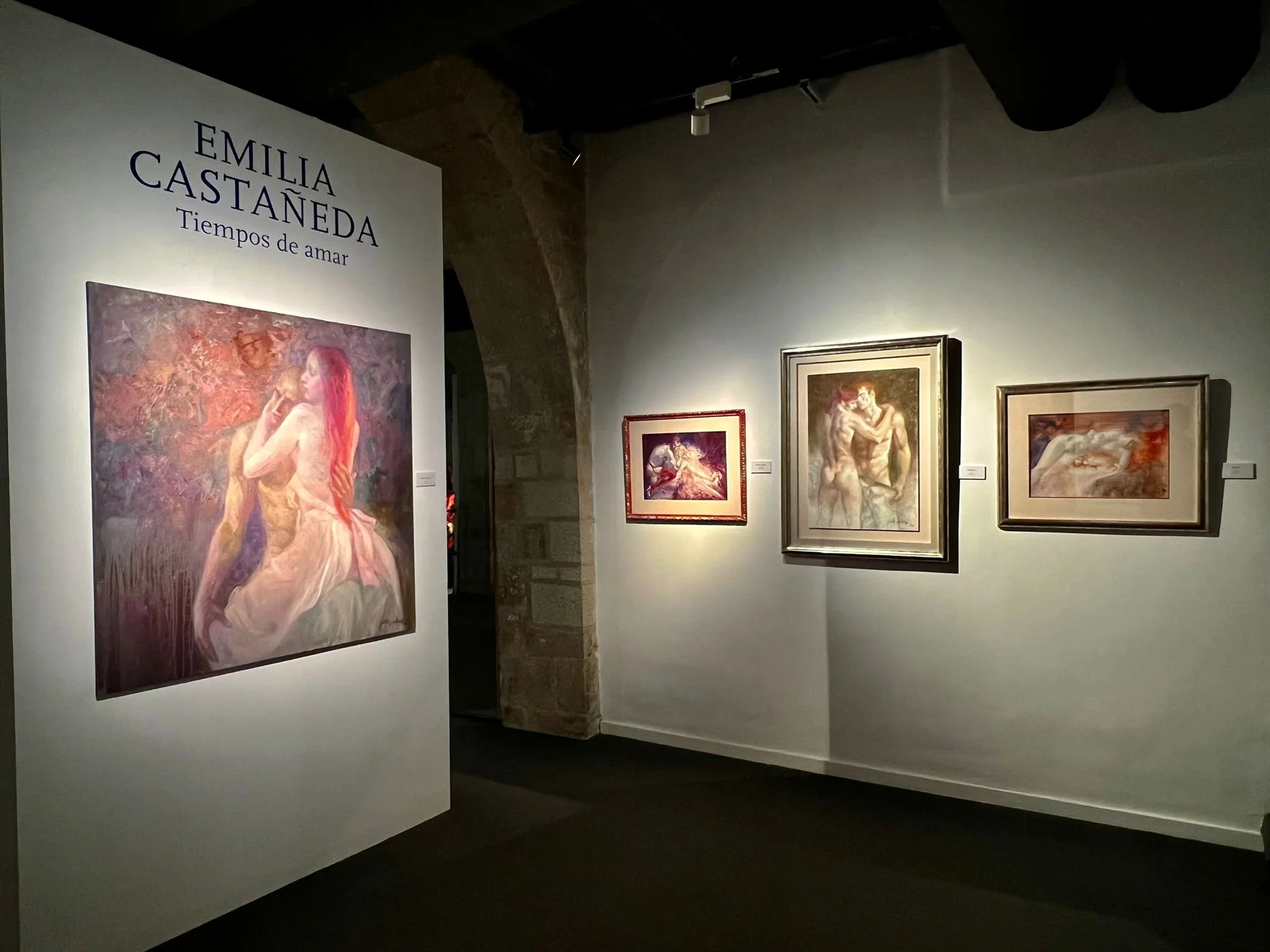 Art exhibition with four paintings displayed on a white gallery wall, featuring abstract and figurative art, with a large poster on the left highlighting Emilia Castañeda's artworks, titled 'Tiempos de amar'.