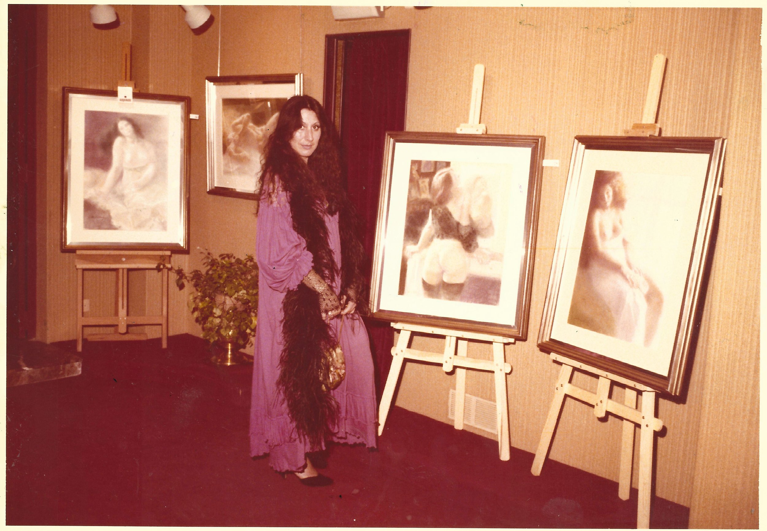 A woman with long curly dark hair, wearing a purple dress and black fishnet gloves, stands in an art gallery surrounded by framed photographs of a woman in various poses on easels. The gallery has beige walls and red carpet flooring.
