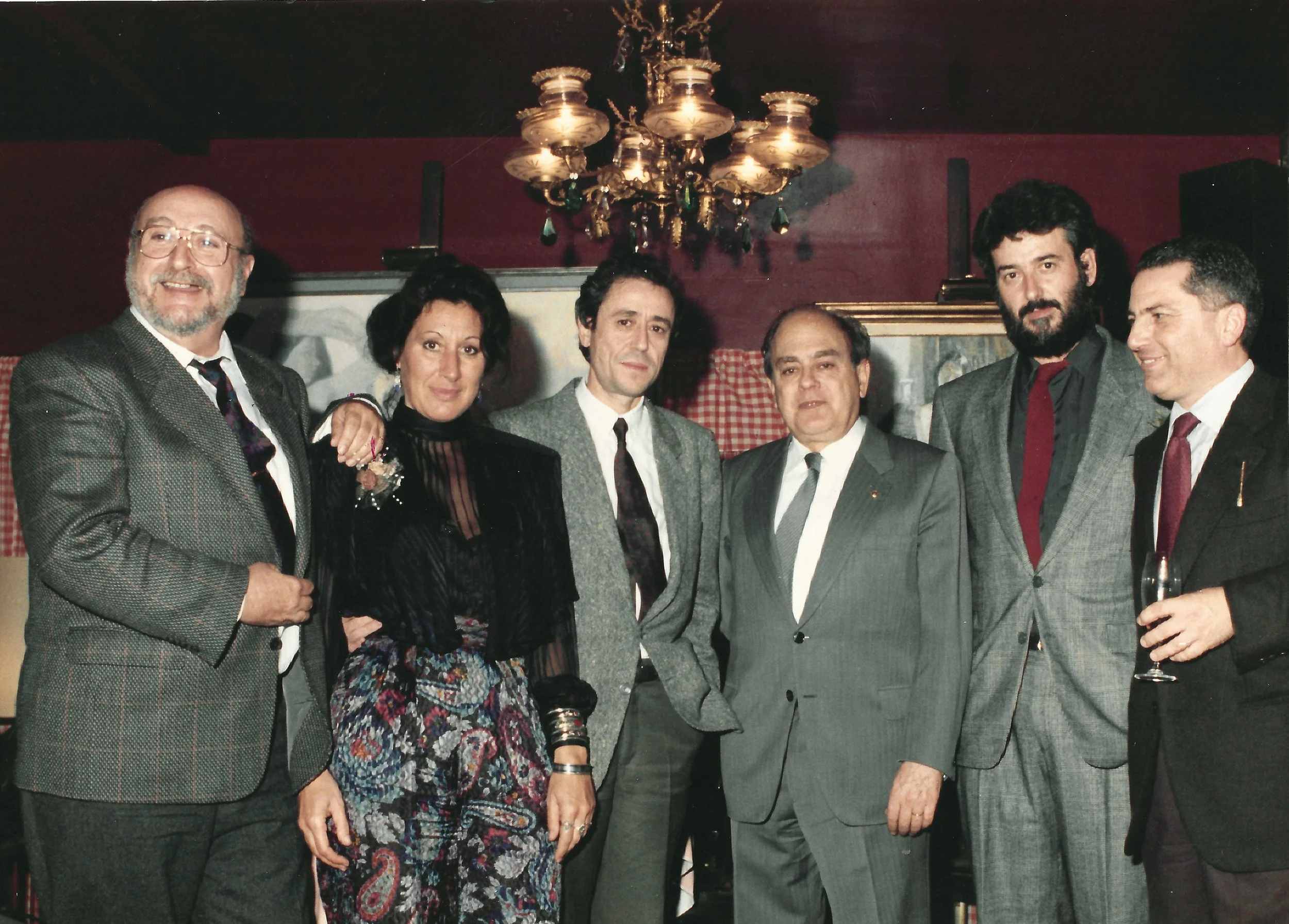A group of seven people, six men and one woman, dressed in formal attire, pose together at an indoor social event. The background features a chandelier and a brick fireplace with a painting above it.