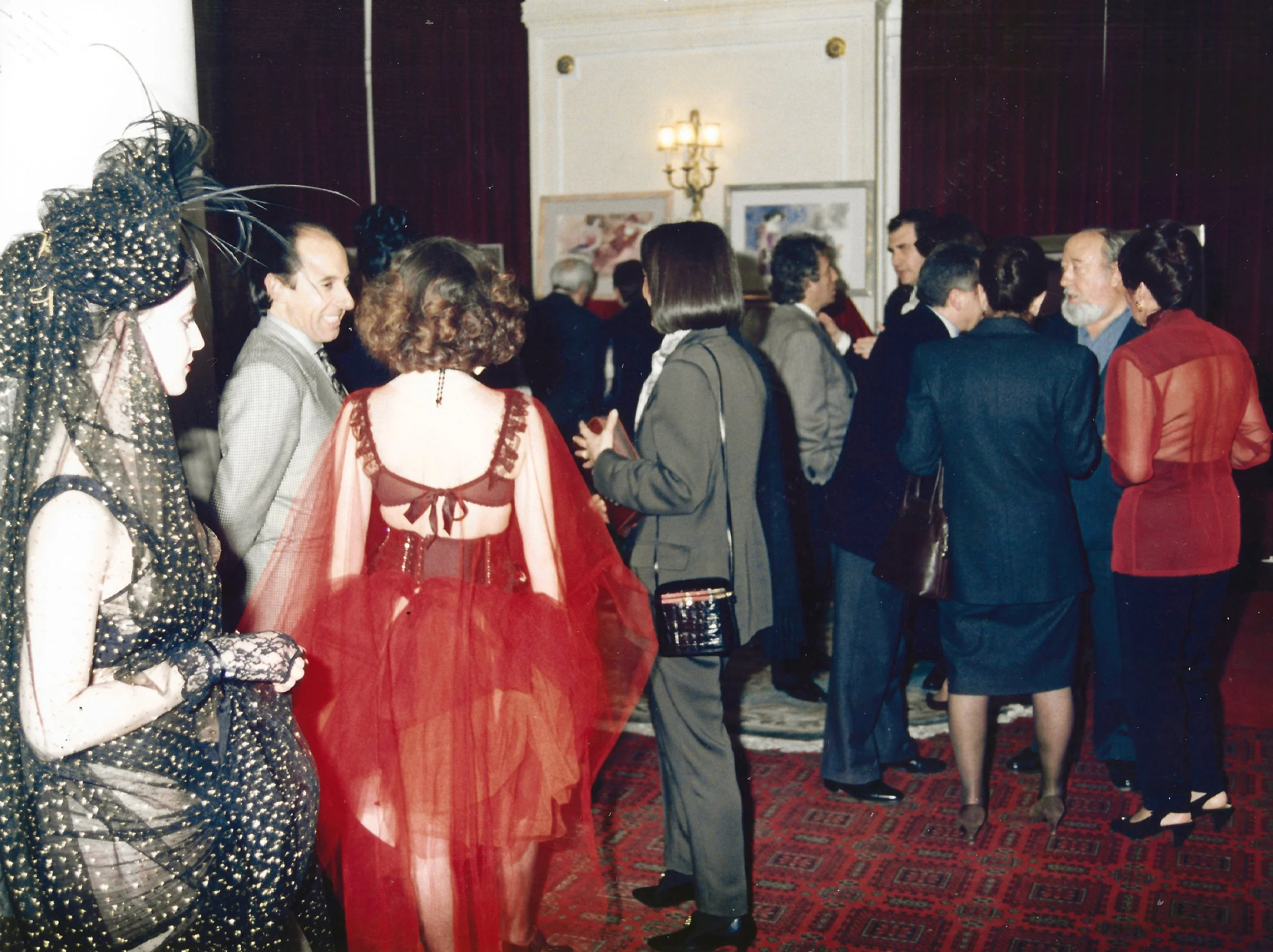 People dressed in formal attire socializing at an event in a room with red curtains, framed artwork, and a patterned carpet.