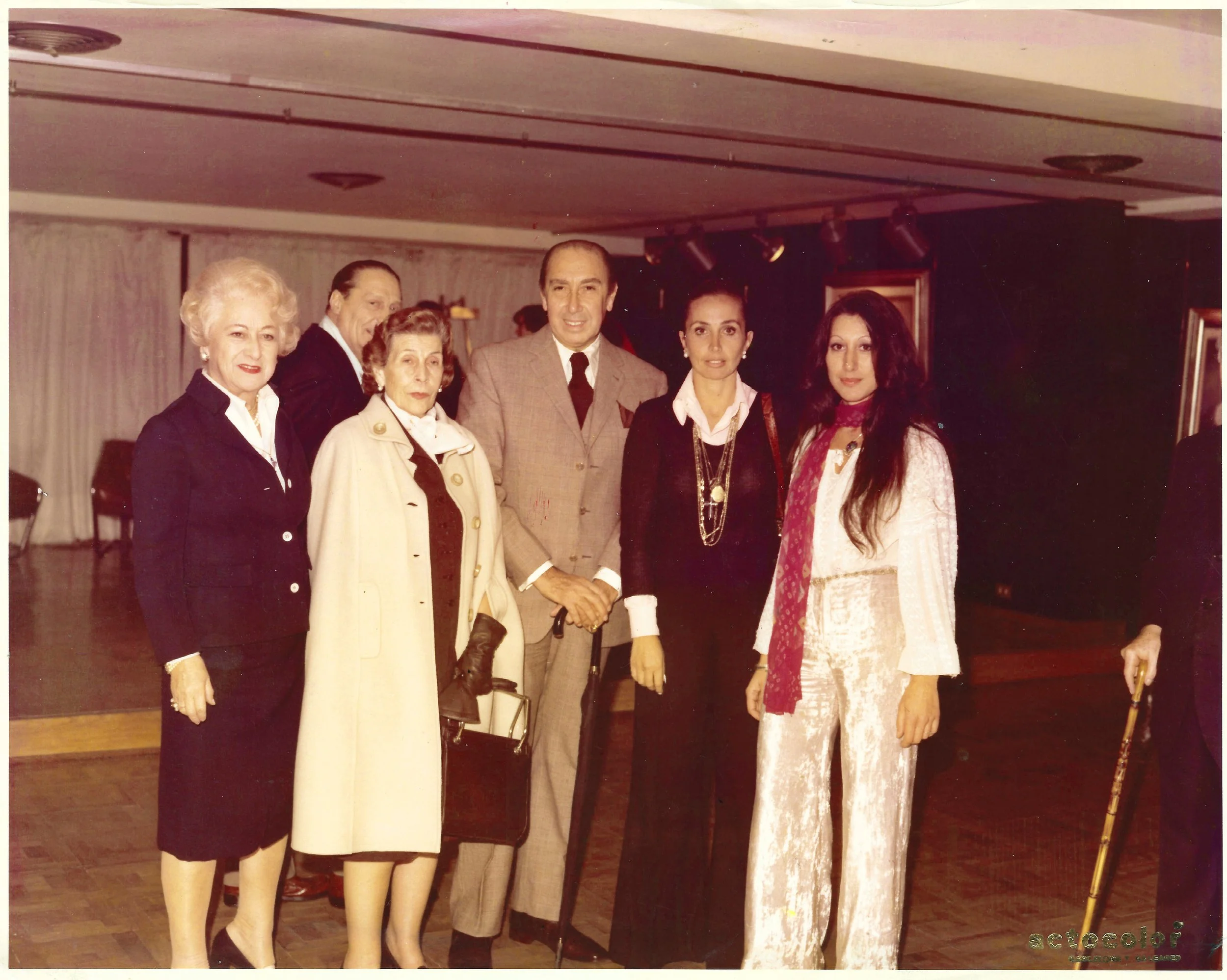 Group of six formally dressed people standing in a room with wooden floors and a dark ceiling, posing for a photo. Four women and two men, some holding walking sticks.