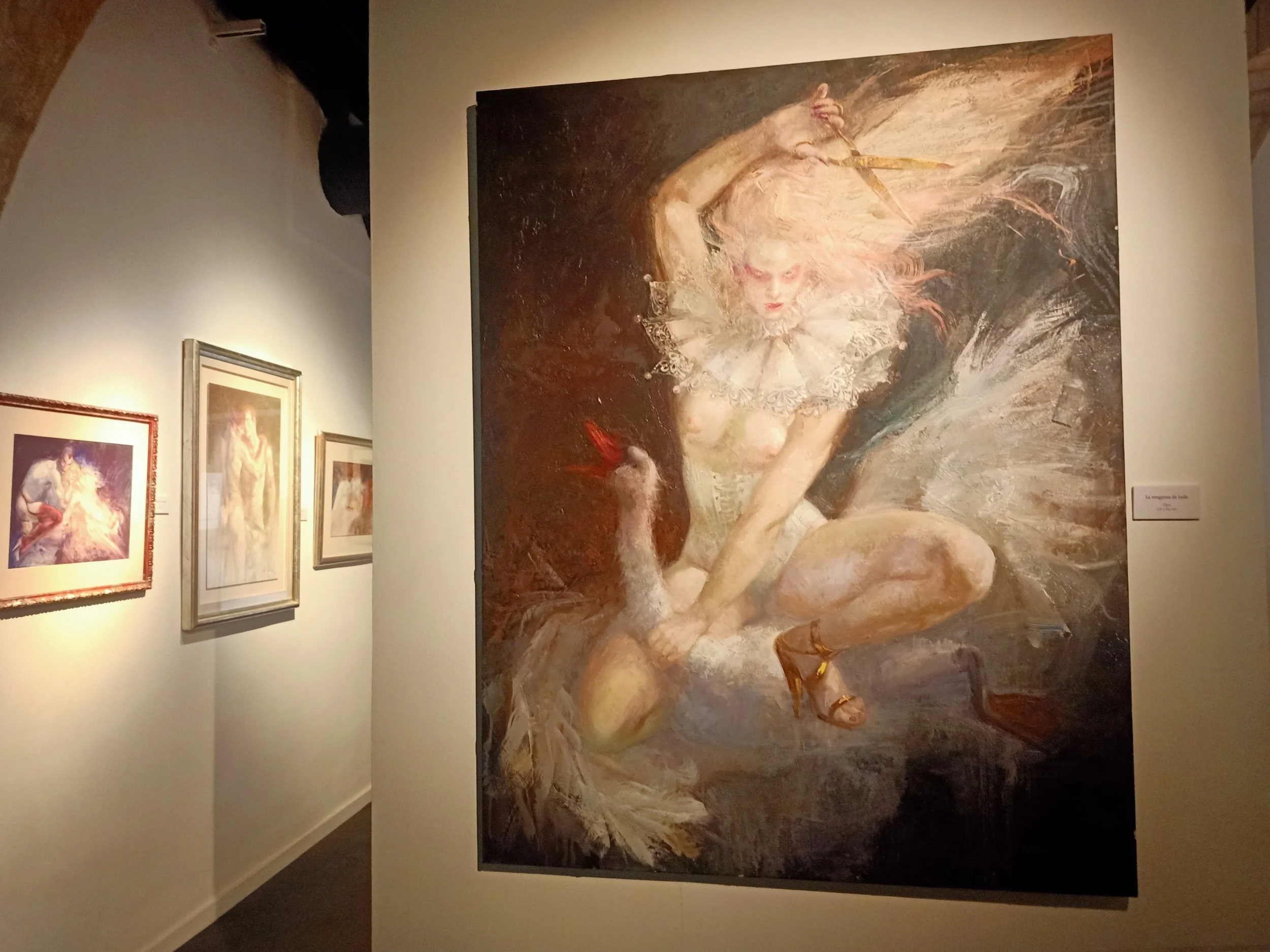 A large impressionist painting of a woman with pale skin and curly hair, wearing a ruffled collar, holding a stick or wand, with a goose on the ground beside her, displayed in an art gallery with other smaller paintings visible on the wall.