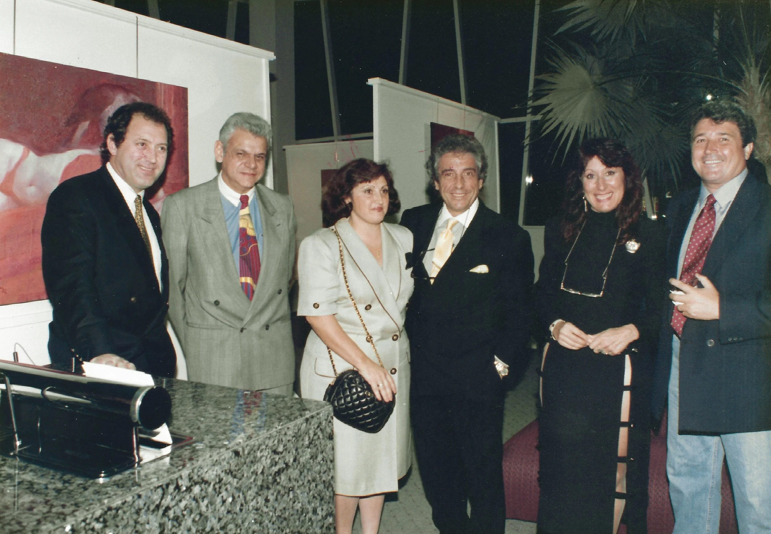 Group of six people standing indoors, dressed in formal and semi-formal attire, smiling at the camera. There are paintings in the background and a granite counter in the foreground.