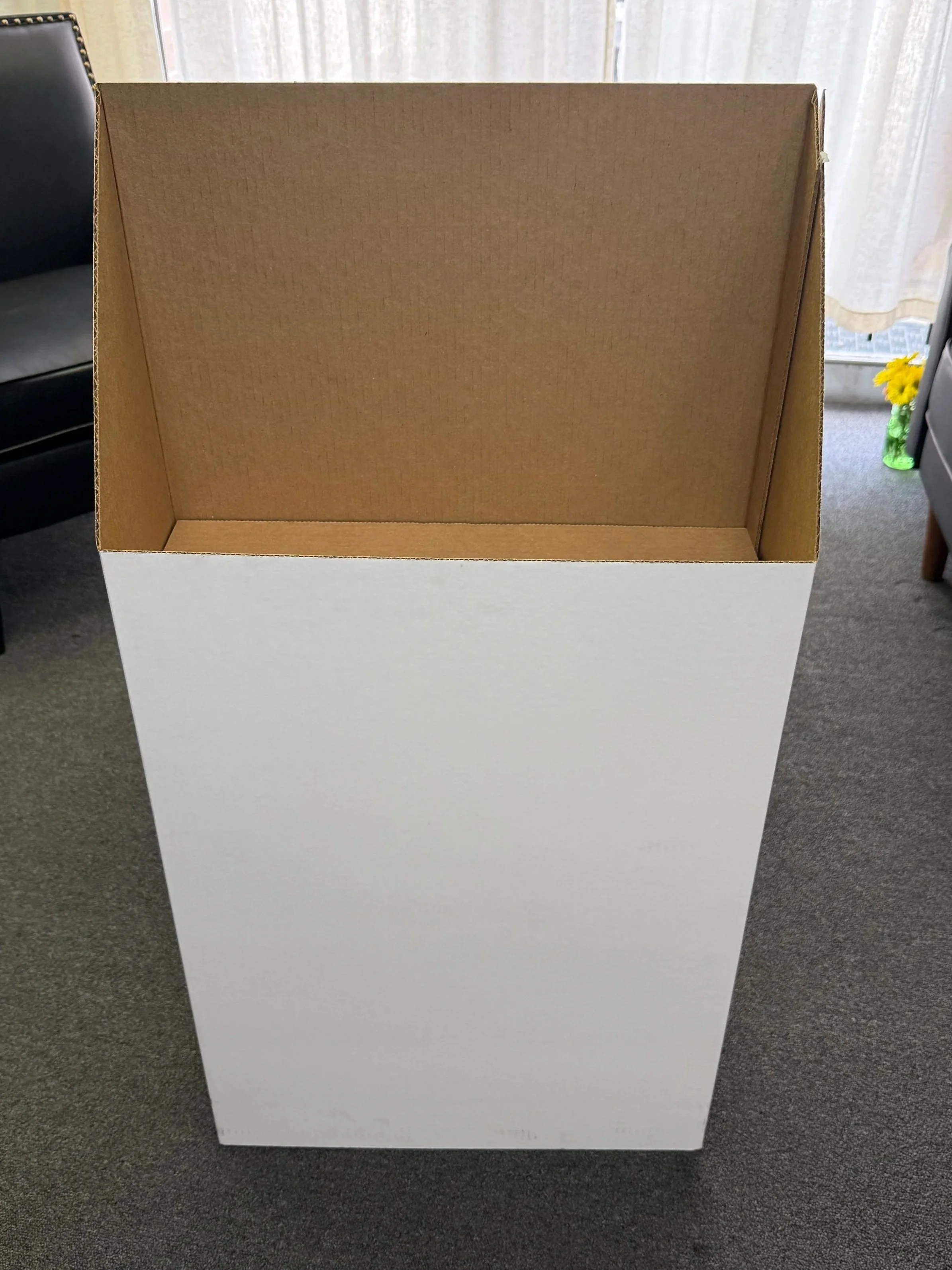 Empty cardboard box on a gray carpeted floor near furniture and a door with a window and a yellow flower in a green vase.