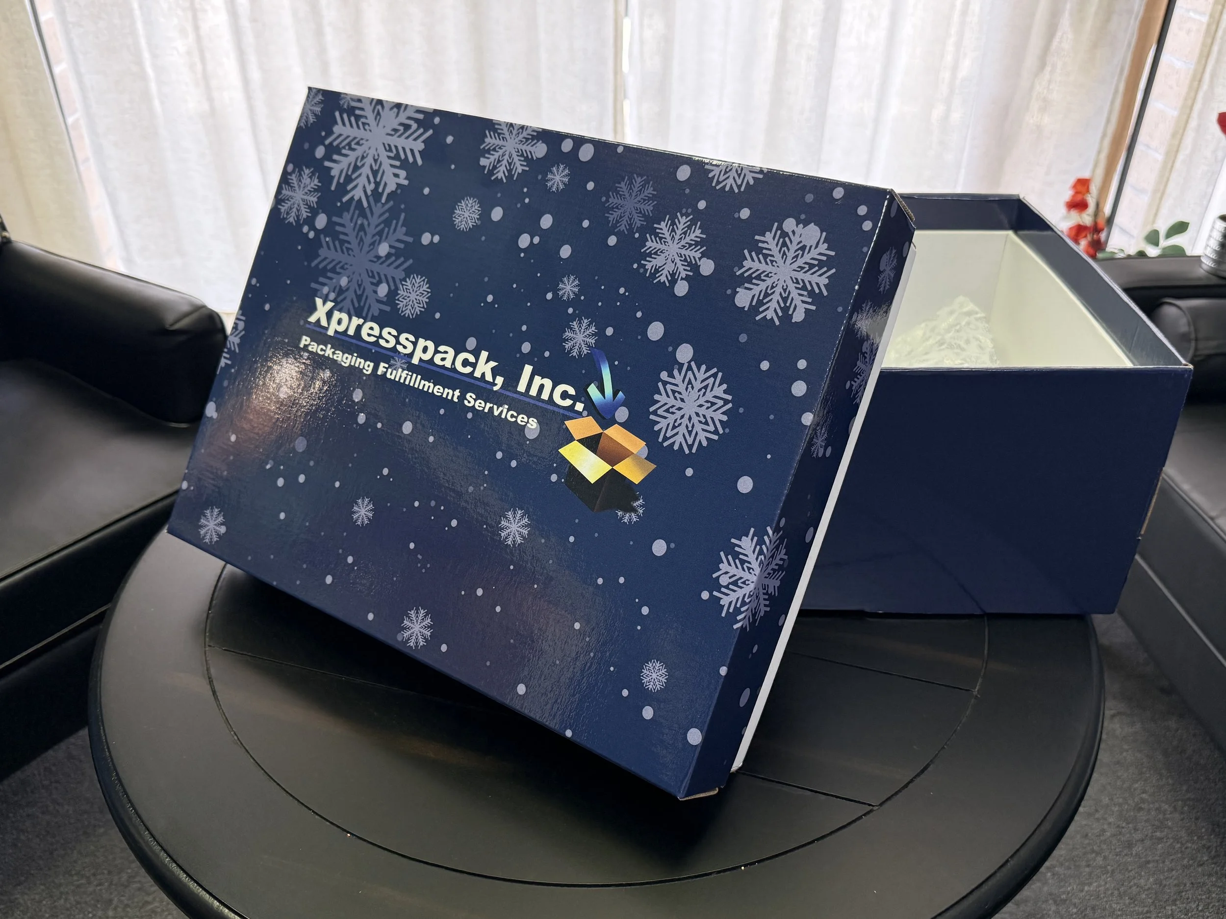 Open blue box with snowflake design, labeled Xpresspack, Inc., Packaging Fulfillment Services, on a black table, with a window and beige curtains in the background.