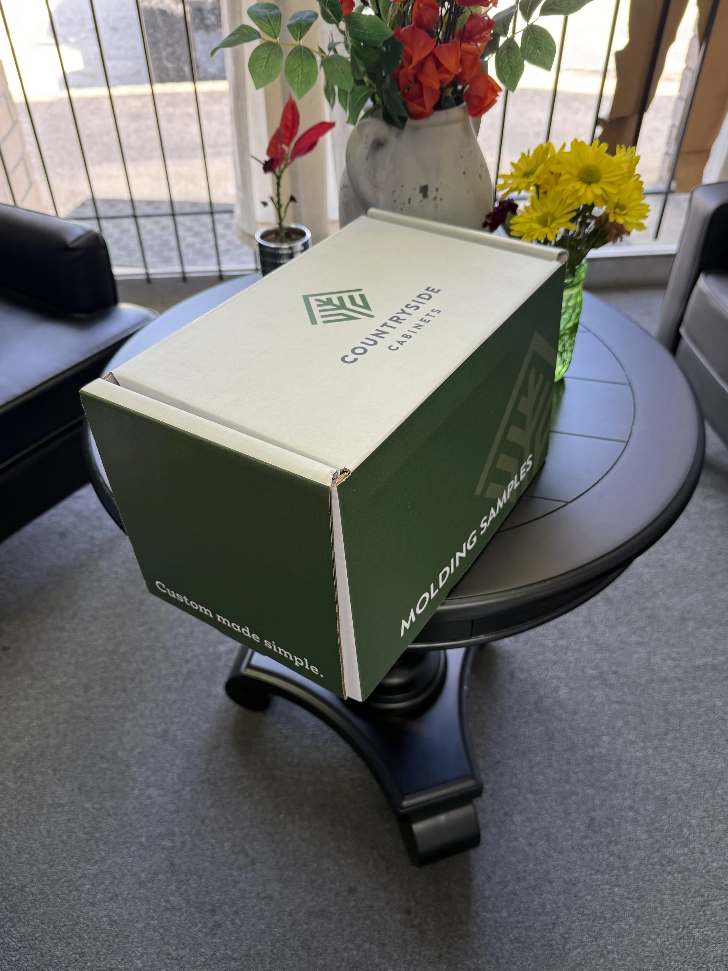 A cardboard box labeled 'Countrieside Cabinets' with additional text 'Molding Samples' and 'Custom made simple' is placed on a round table with a vase of yellow flowers and another vase of flowers in the background, near a window with a view outside.
